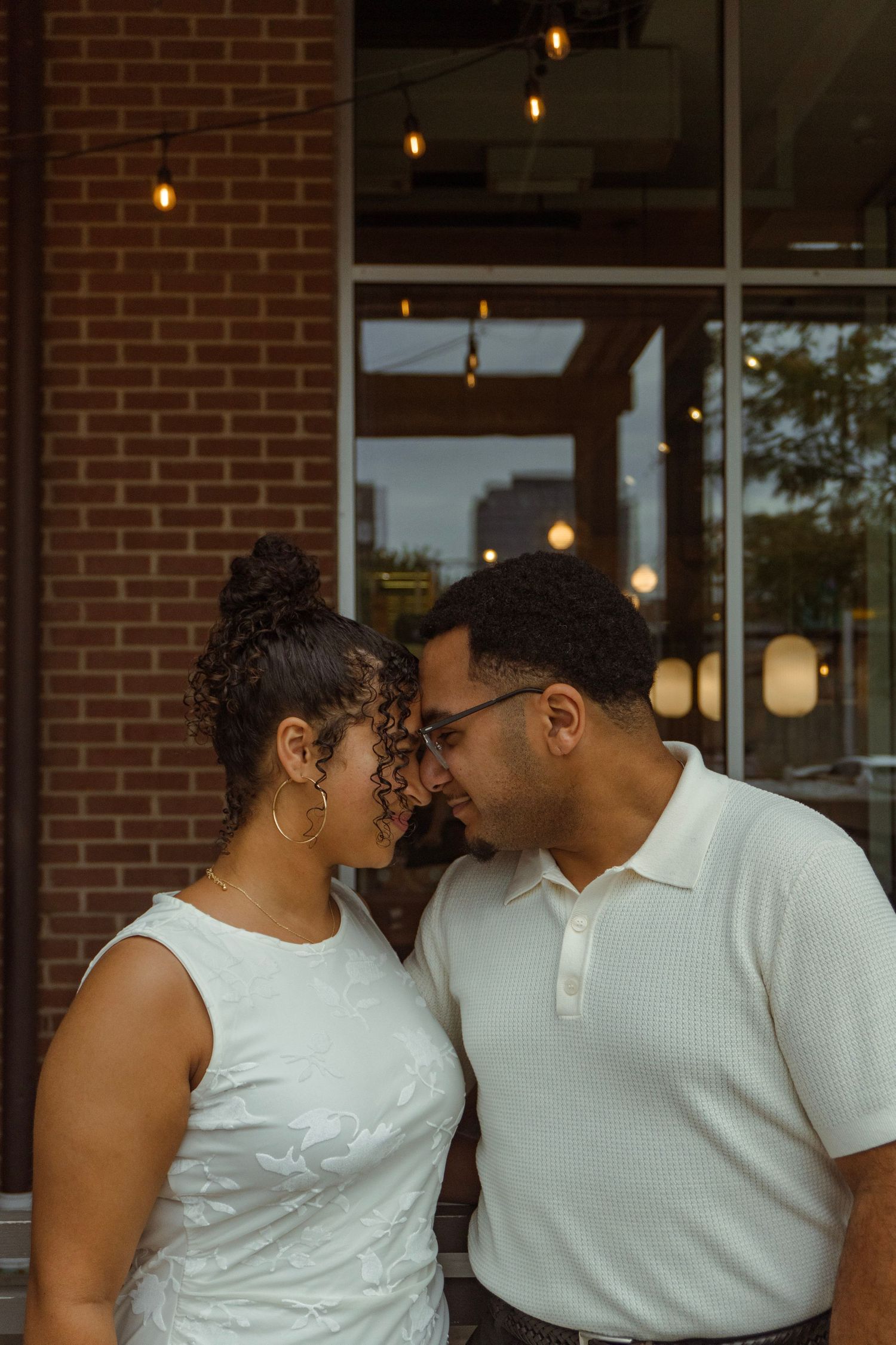 A couple shares a romantic forehead touch outside a brick building with warm string lights in the background.