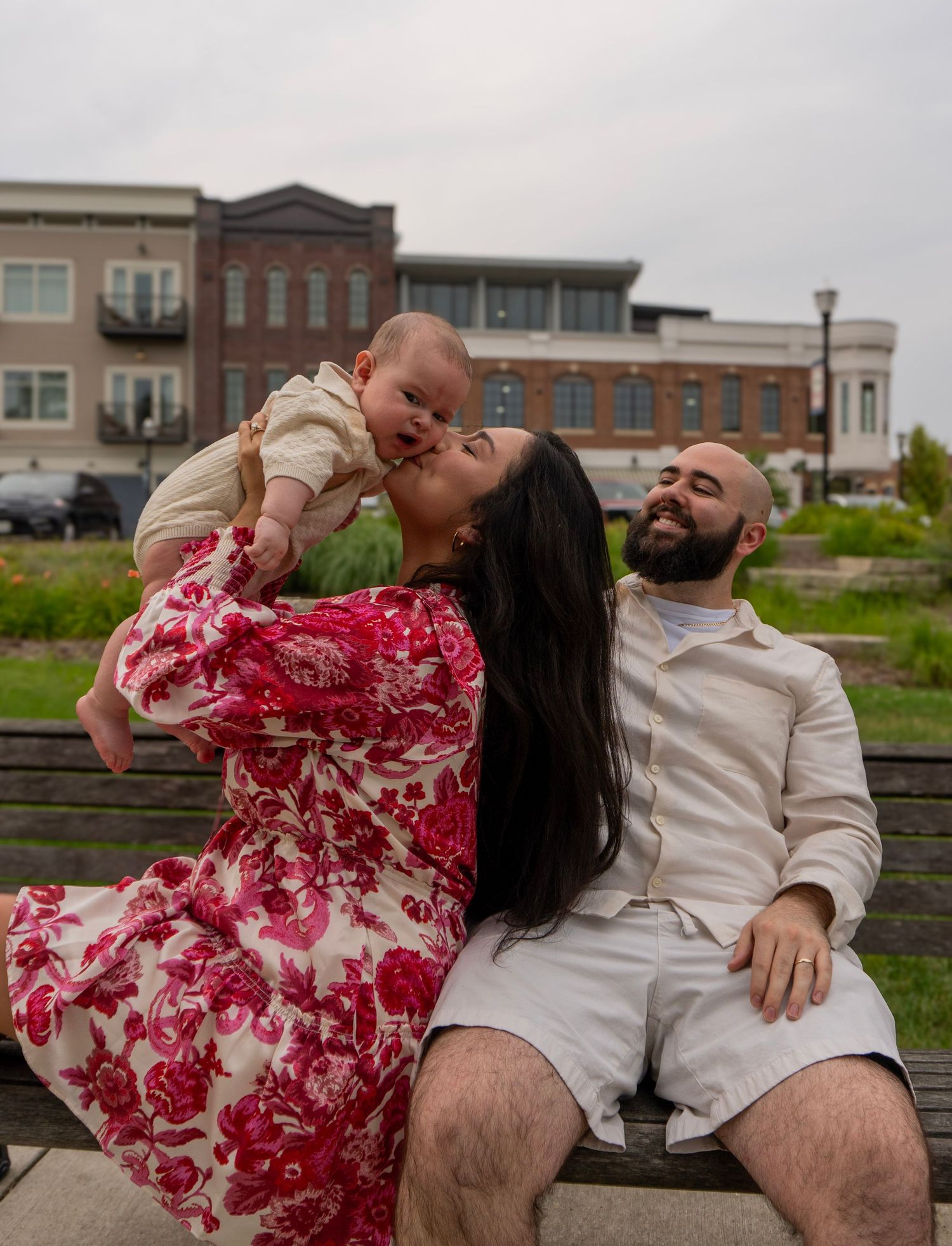 A loving mother in red floral dress kissing her baby on a park bench beside her laughing bearded husband.