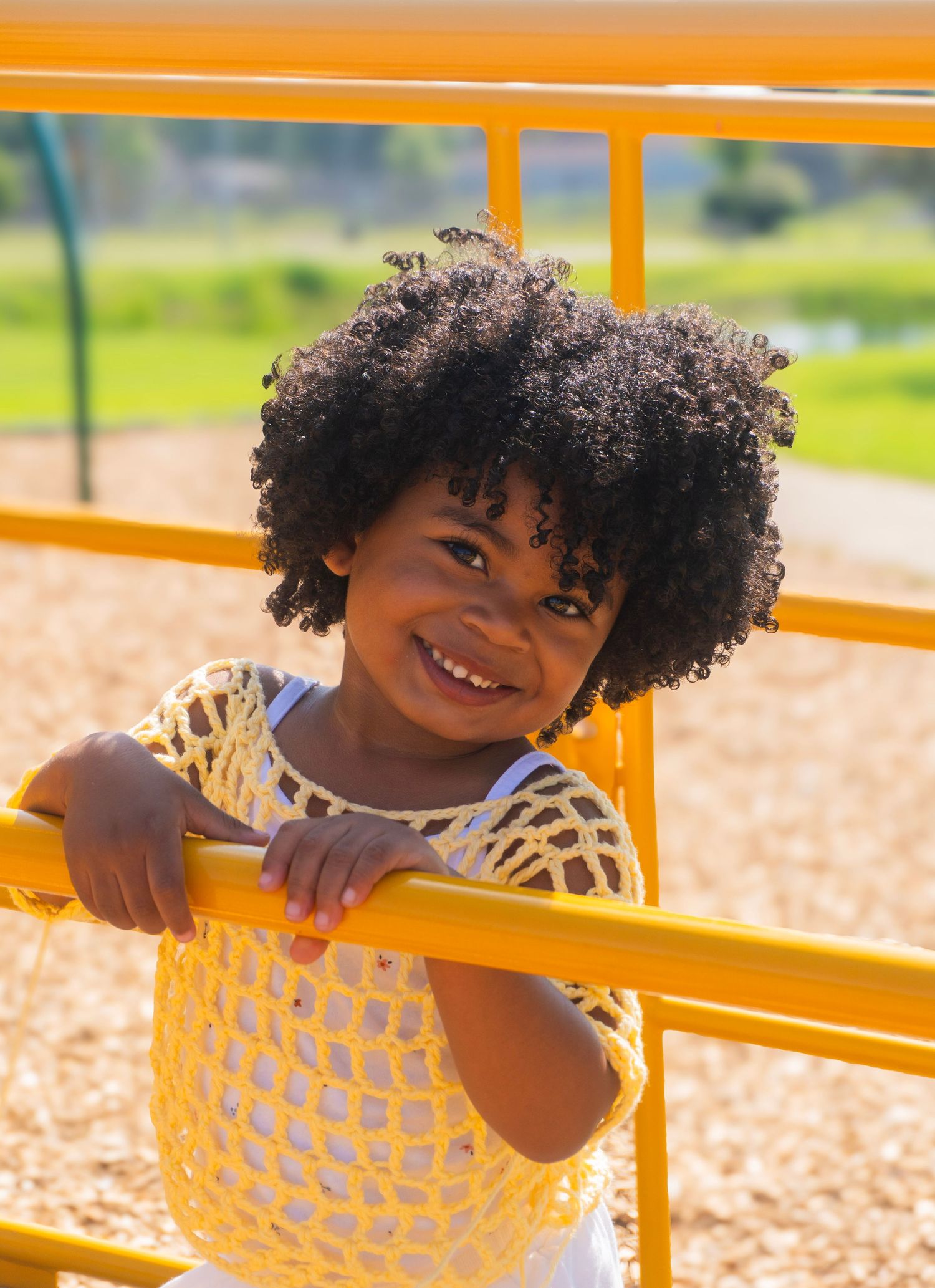 A smiling Black child with a natural afro holds a yellow playground bar wearing a yellow crochet top.