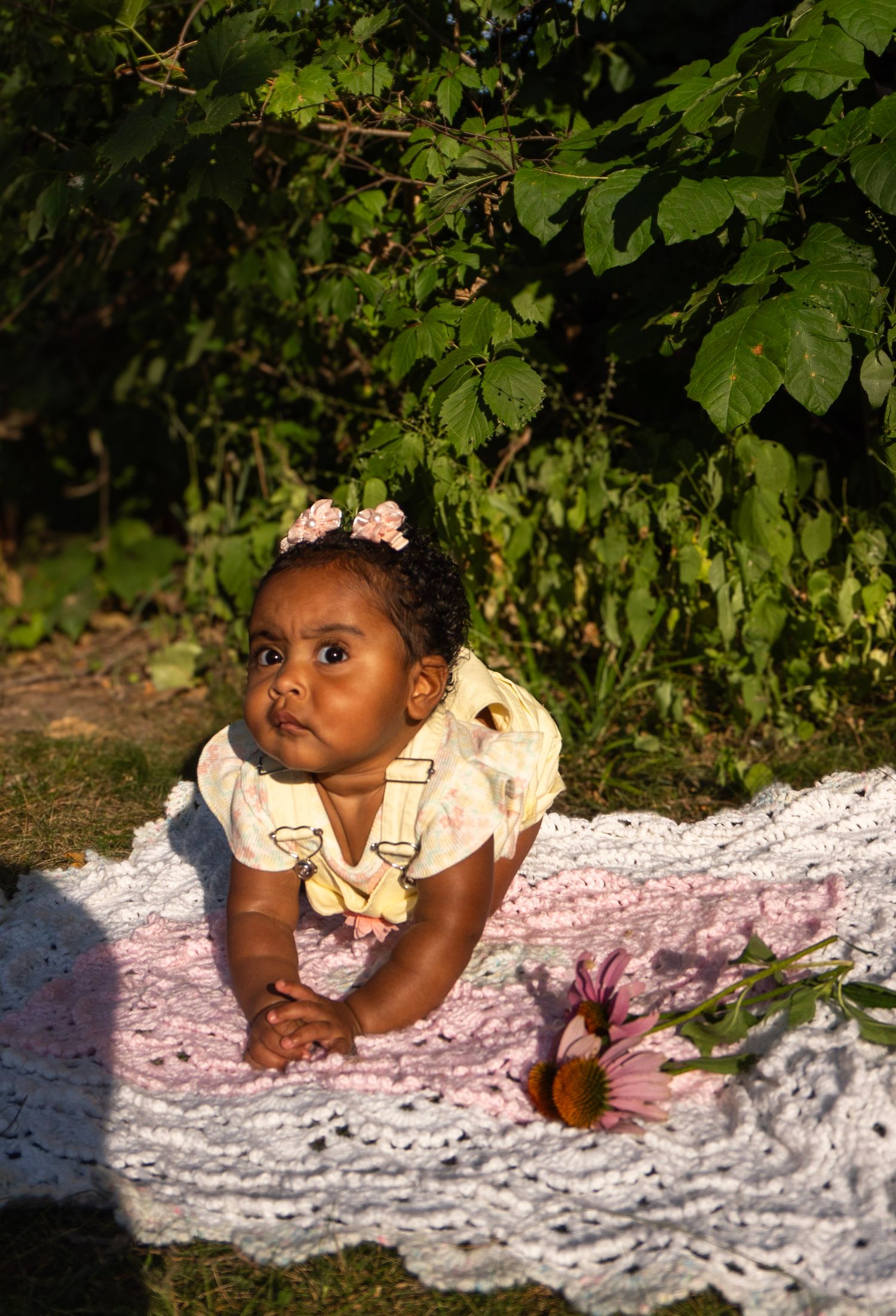 A baby girl in a yellow floral outfit props herself up on a pink crochet blanket outdoors.