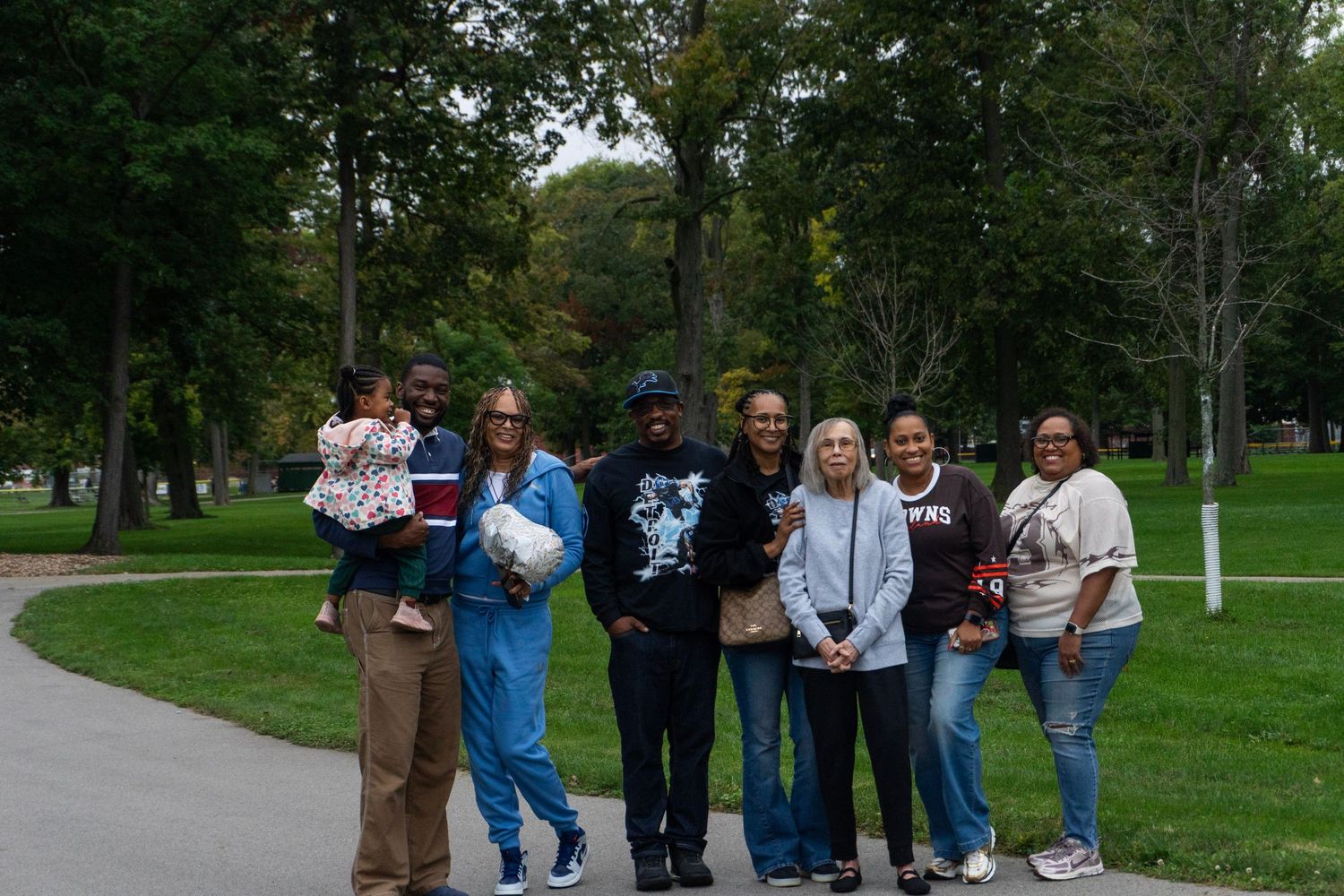 Group of seven people posing together on a park path surrounded by green trees on a cloudy day.