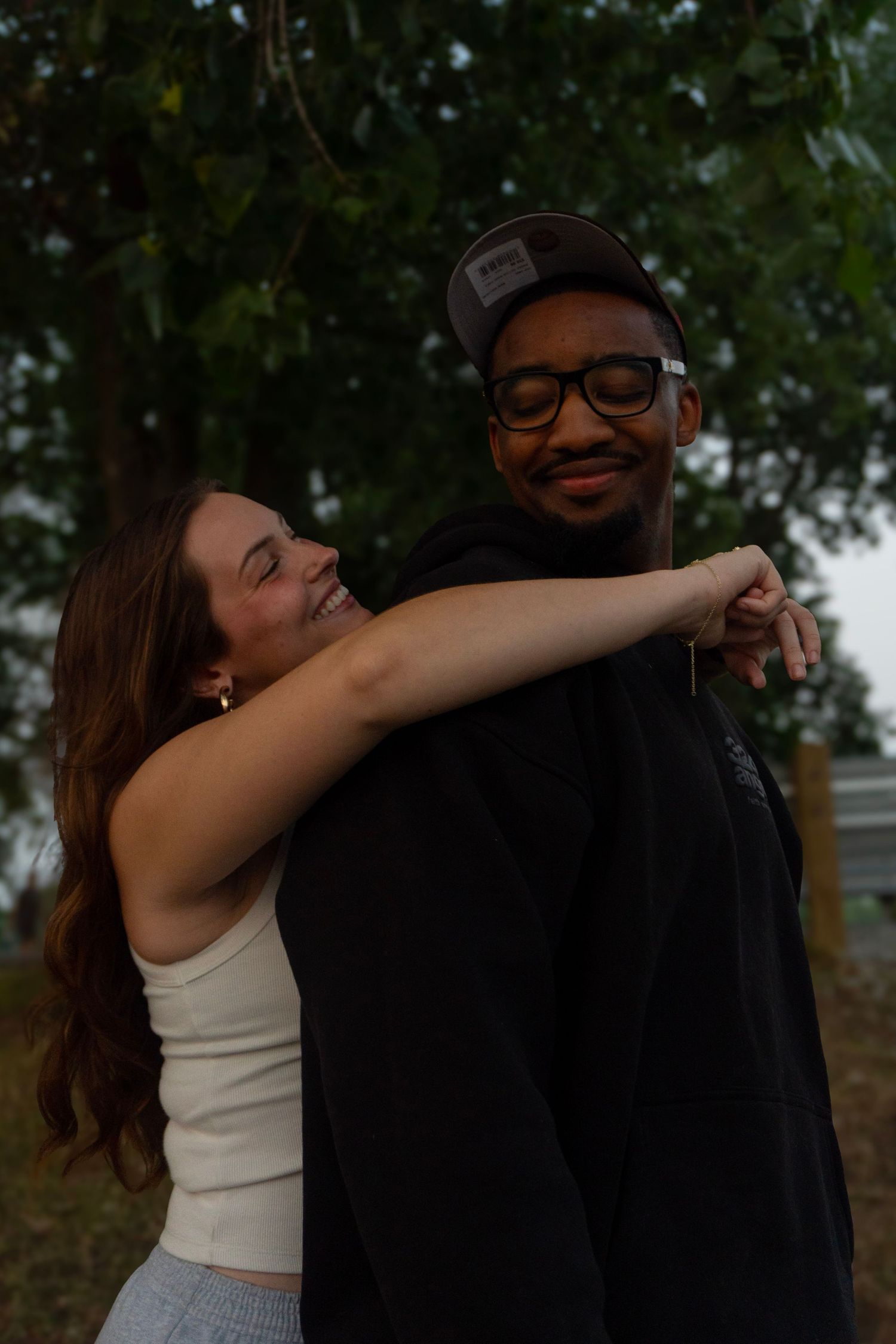 Woman hugging man from behind while smiling outdoors near large green tree at dusk.
