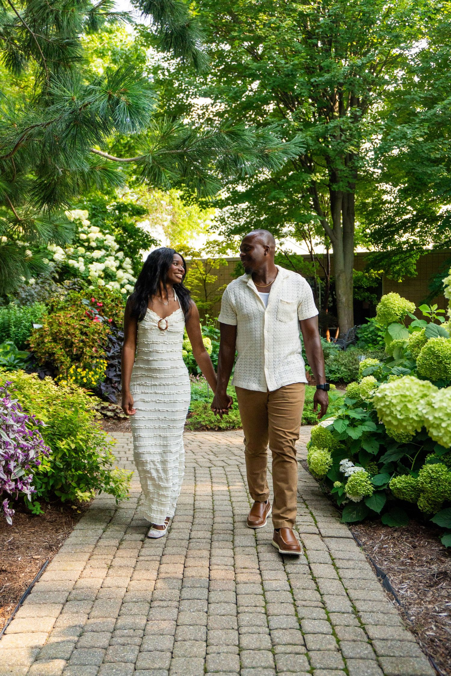 A couple holds hands while walking along a brick garden path surrounded by lush greenery and hydrangeas.