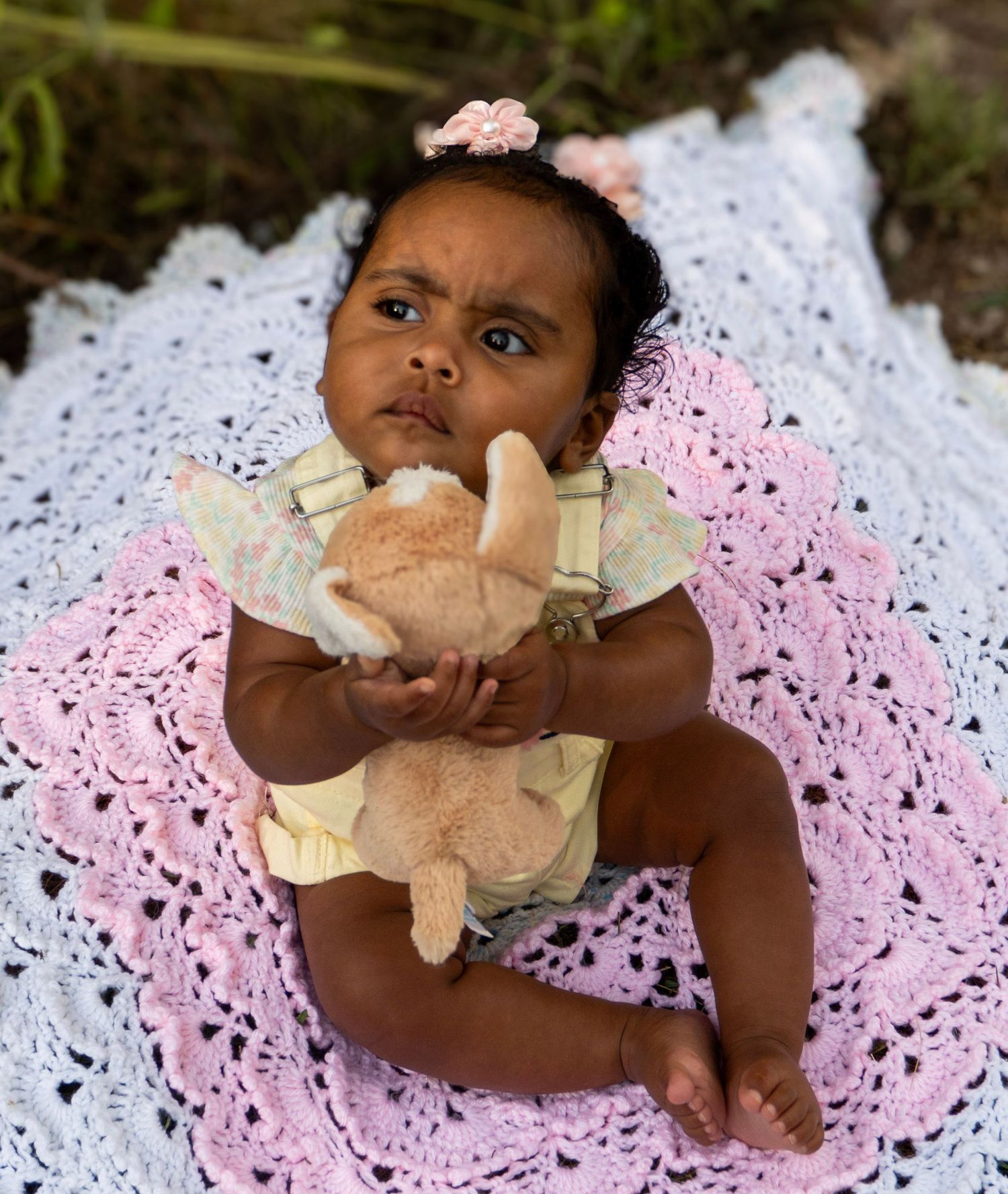 Adorable Black baby girl in yellow overalls holds stuffed bunny toy while lying on pink crochet blanket.