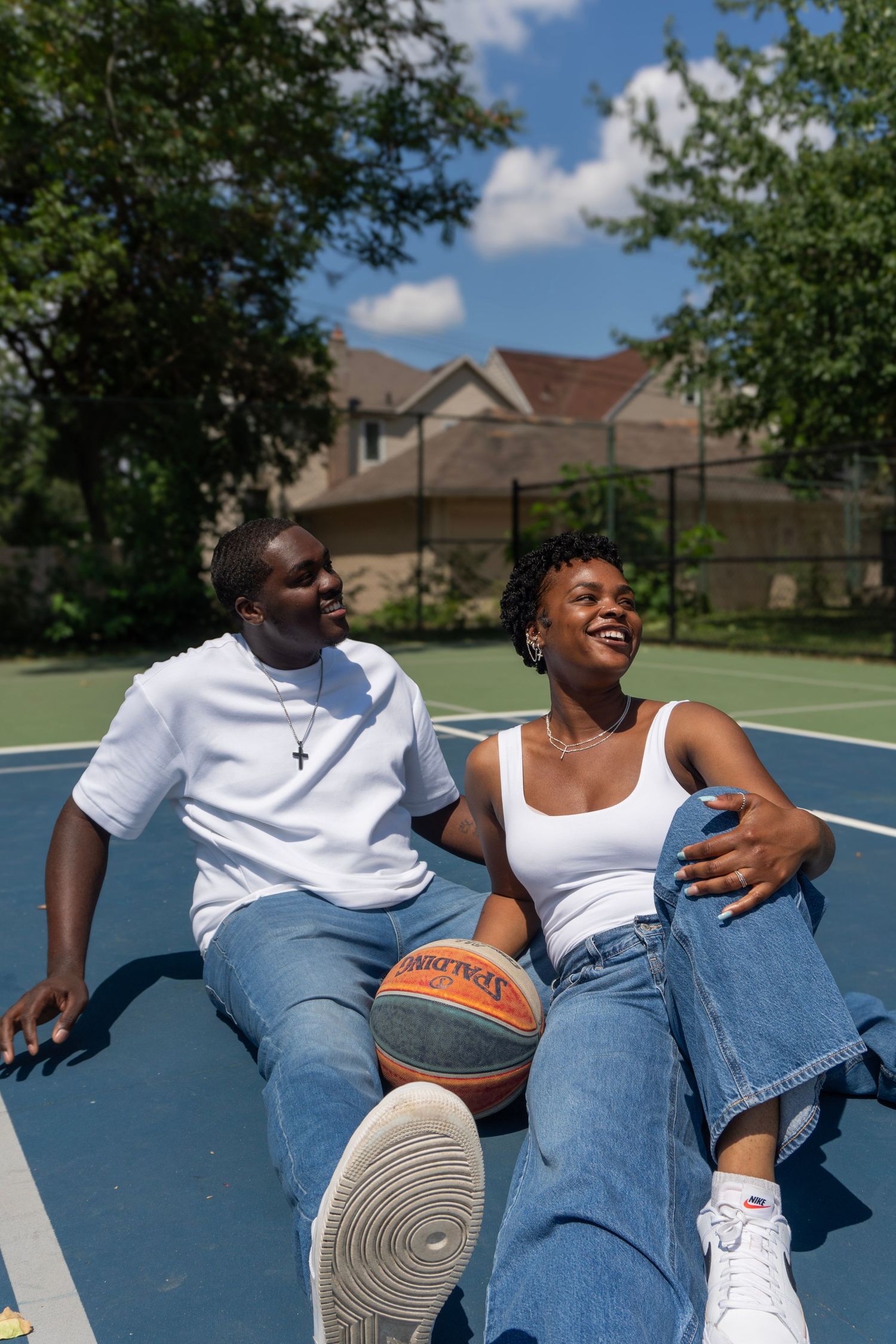 A happy Black couple sits closely together on a blue outdoor basketball court with a basketball.