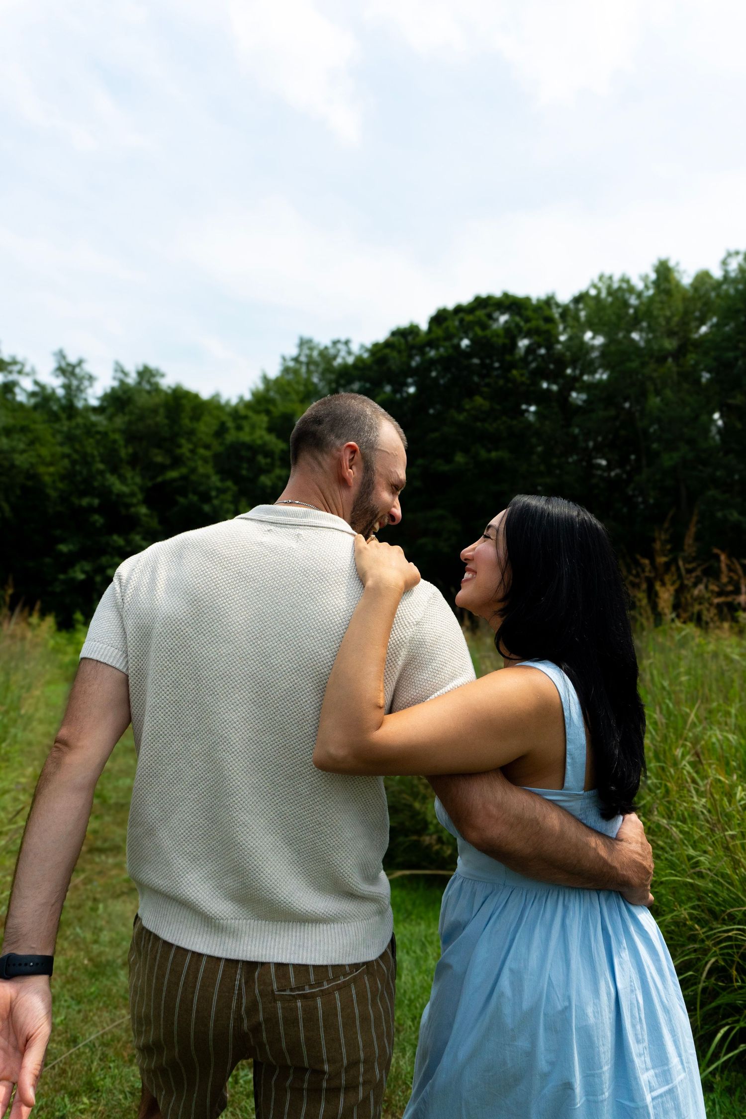 Couple embracing outdoors in green field, woman in blue dress laughing with man in gray shirt.