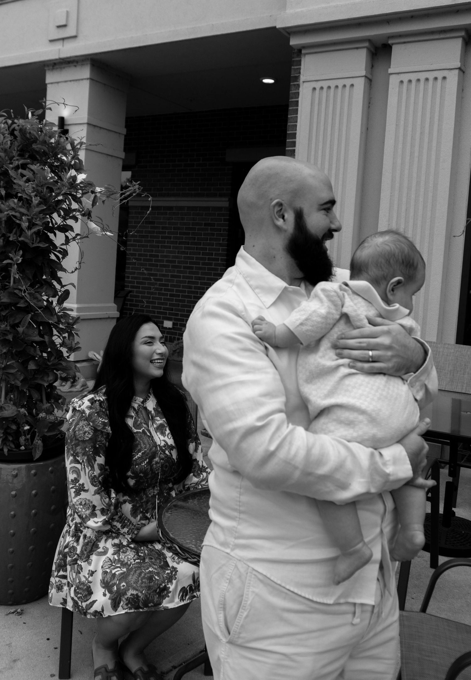 Black and white portrait of bald bearded father tenderly holding infant while mother smiles in background.