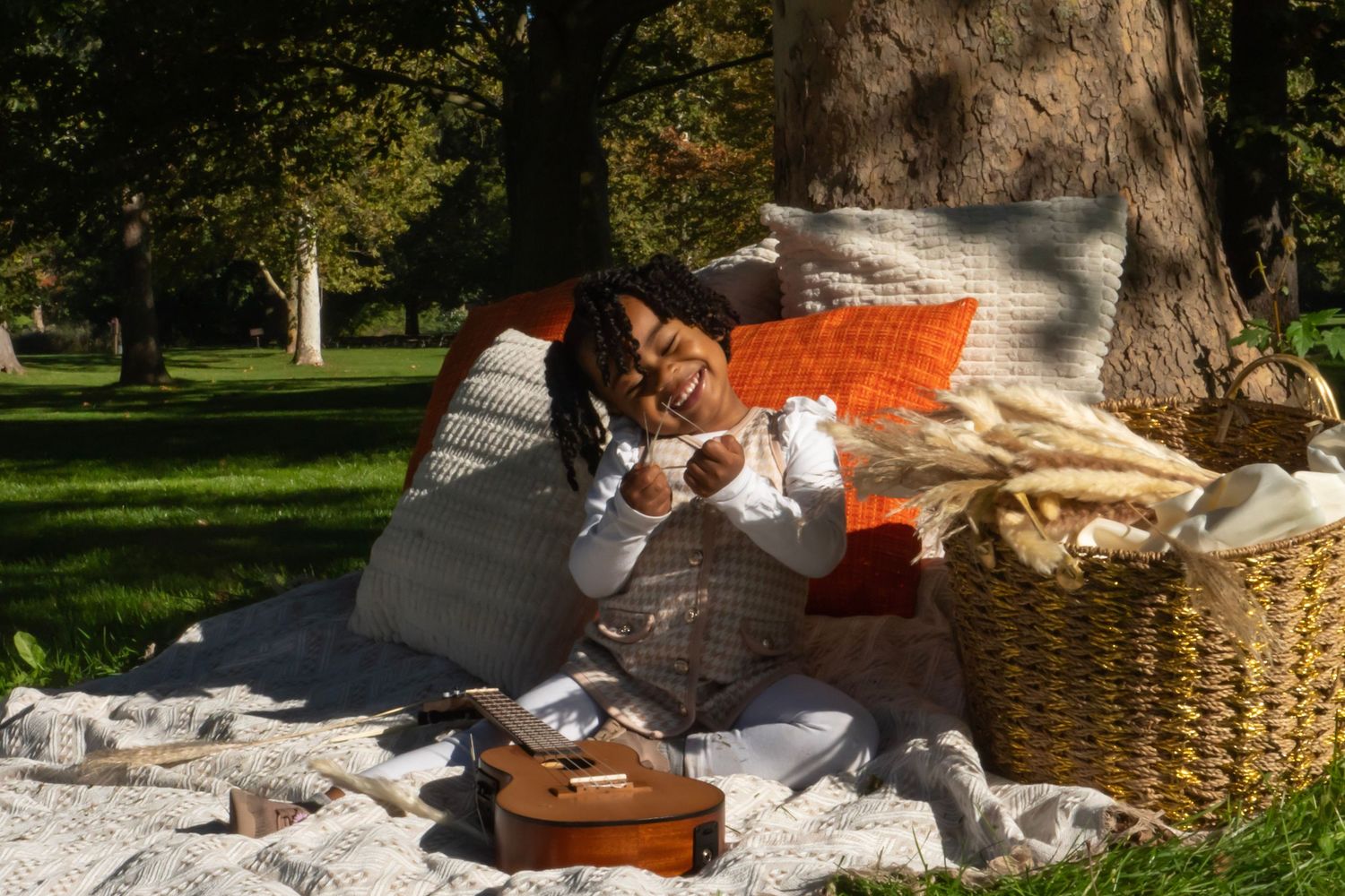 Woman relaxing on a picnic blanket in a park, leaning against pillows near a wicker basket and ukulele.