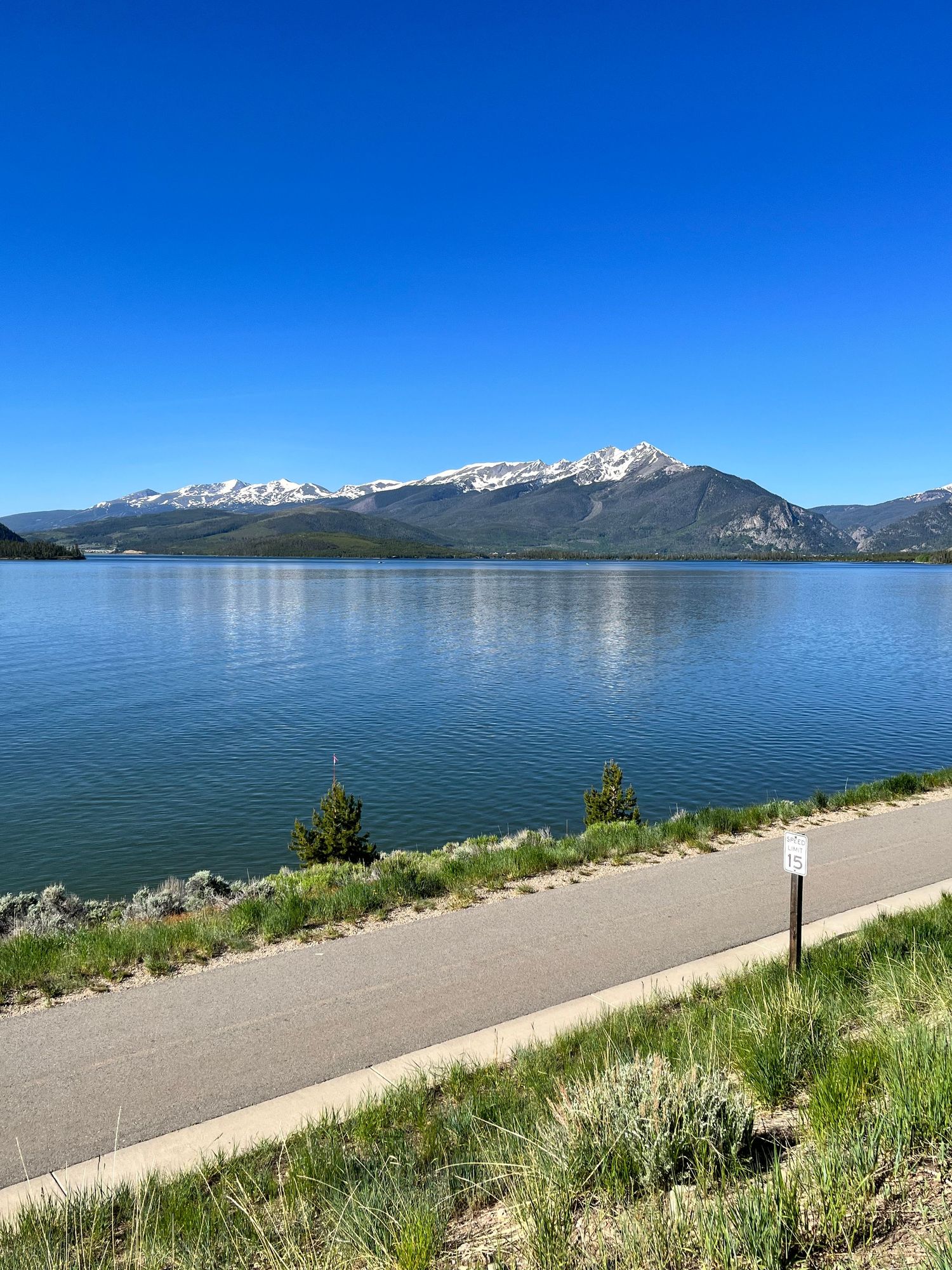 A large body of water surrounded by mountains, a bright blue sky, and grass.