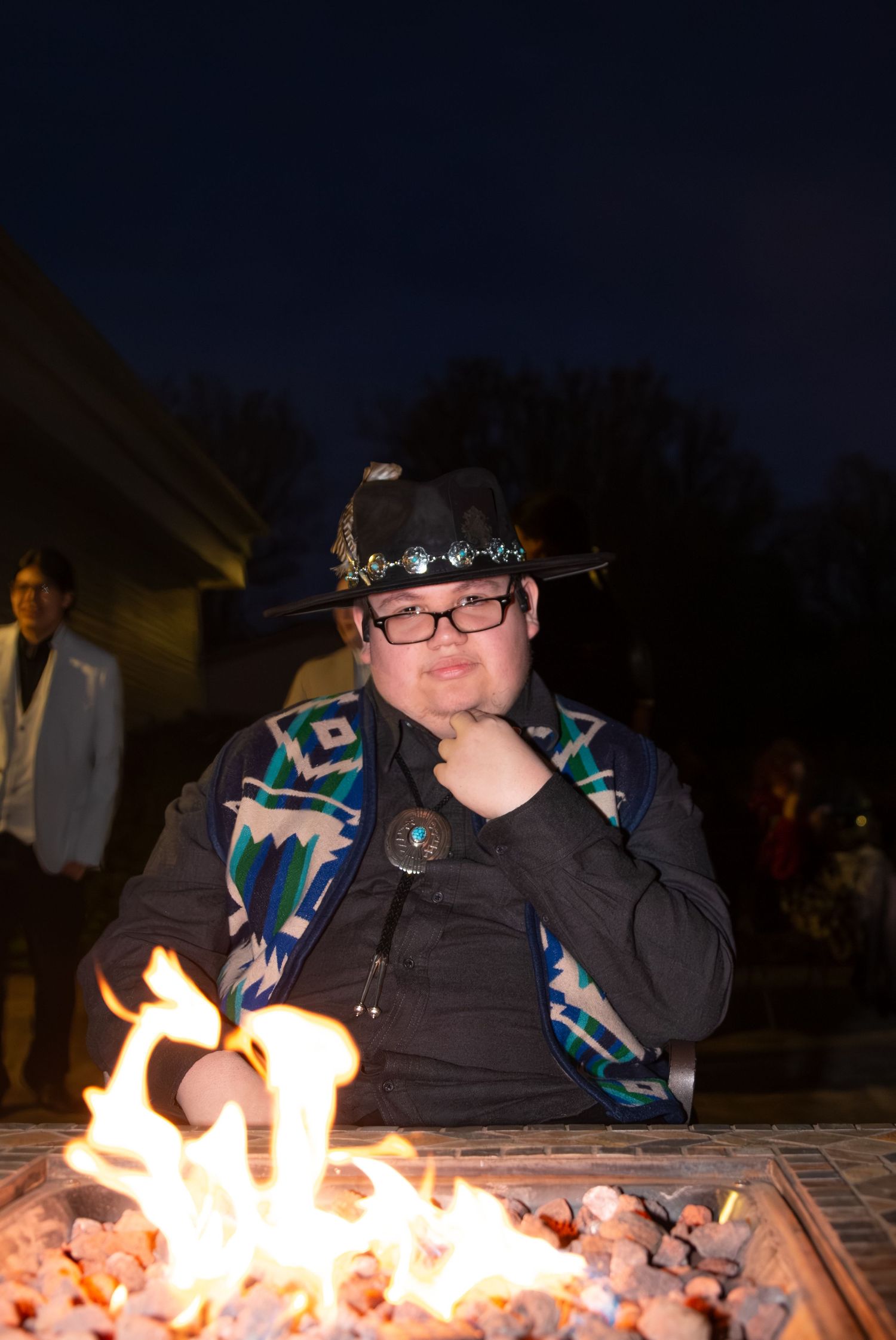 A person looking into a camera in front of a fire, thinking with their hand on their chin.