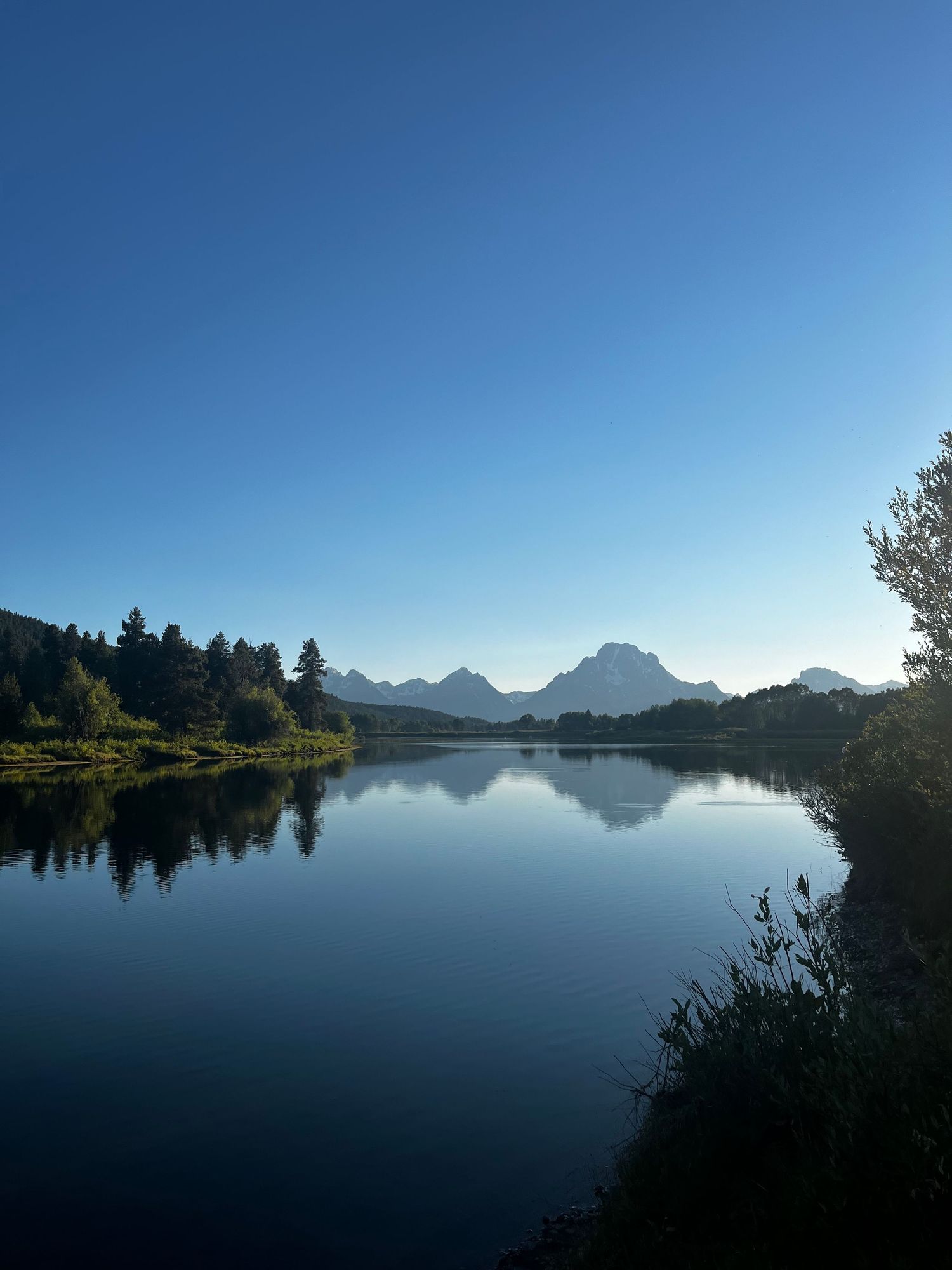 A large body of water surrounded by varying levels of grass and trees, with mountains and a bright blue sky gradient above it.