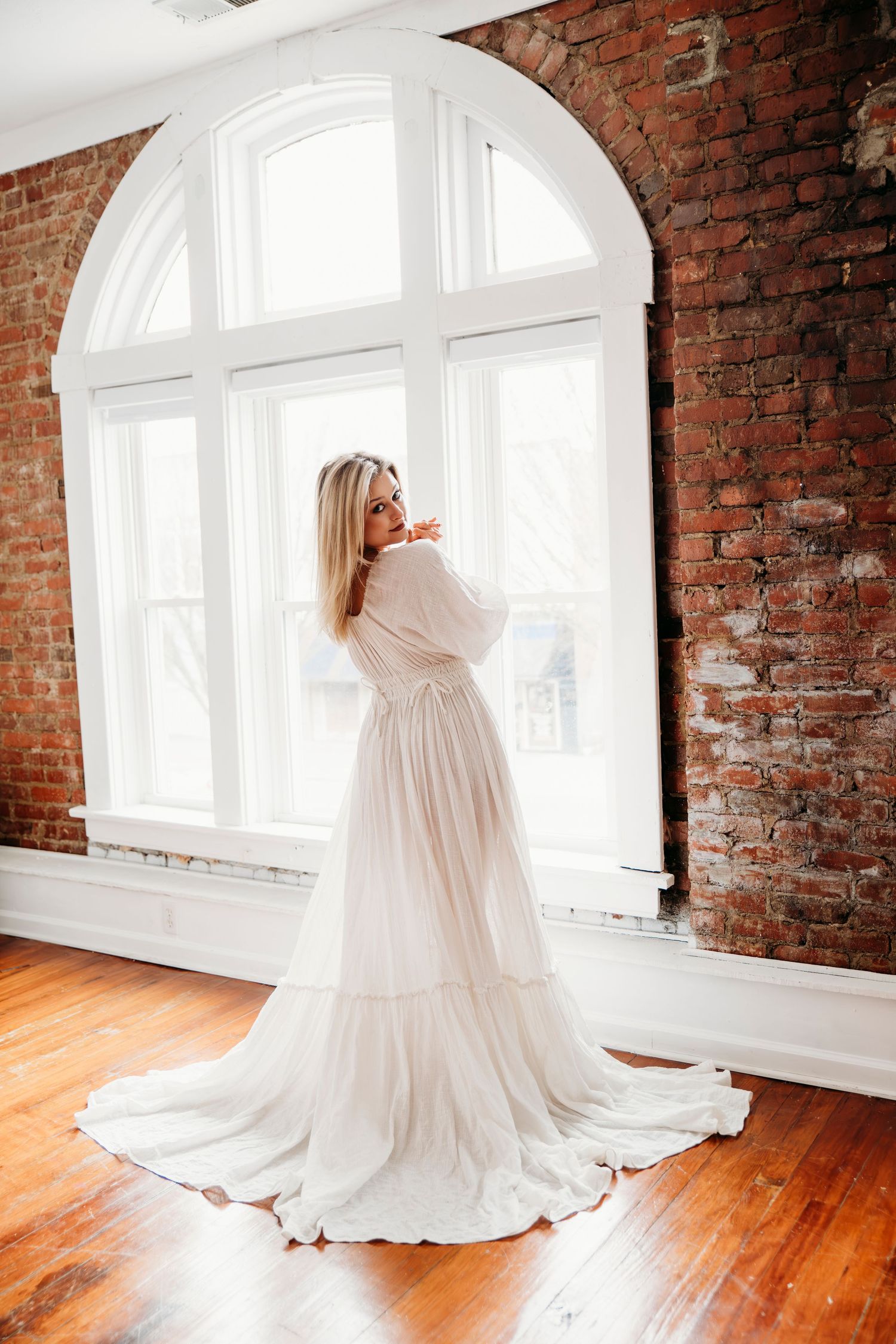 Elegant white wedding dress with flowing train displayed in rustic brick room with arched windows and hardwood floors.