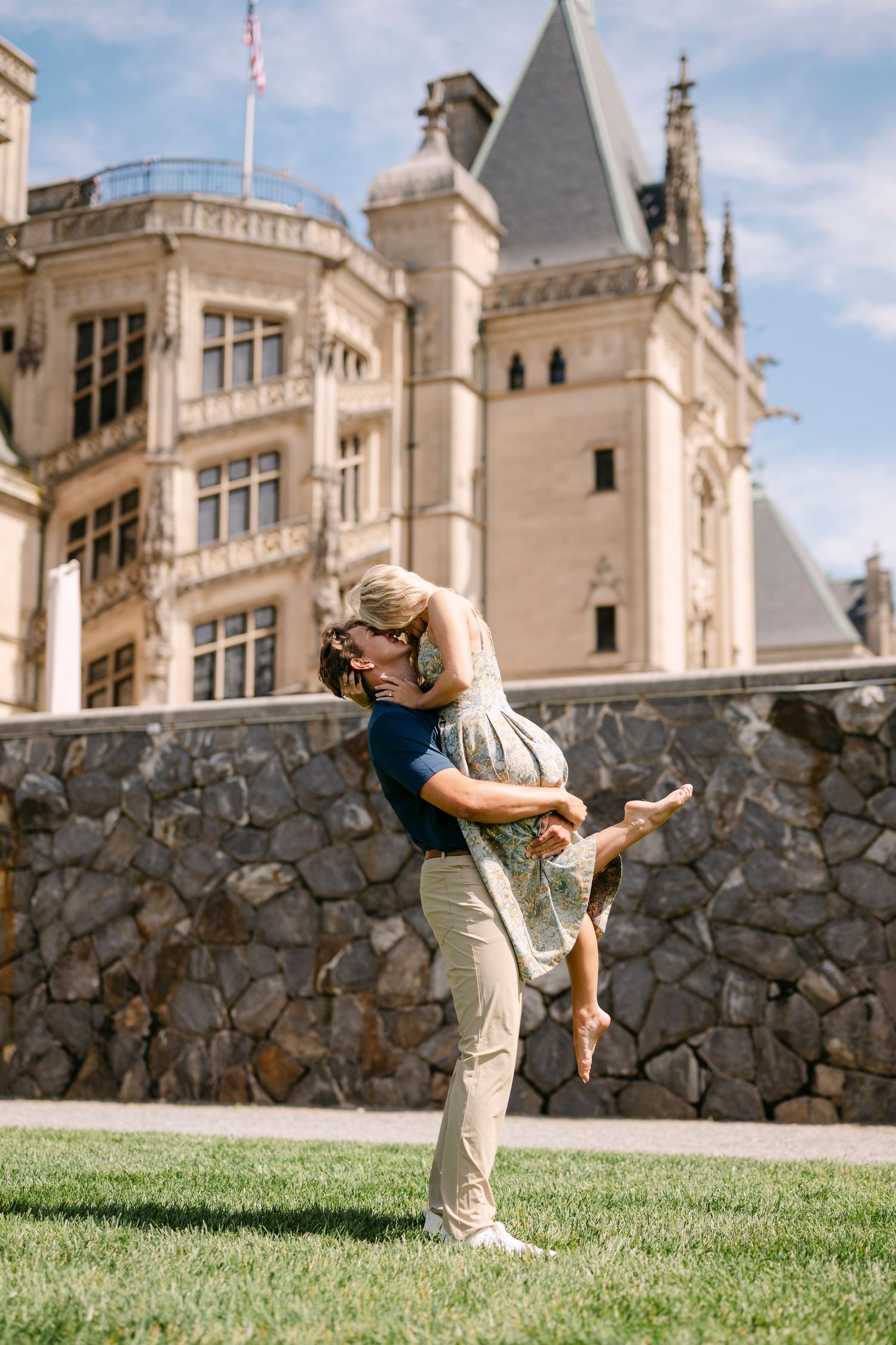 Couple embracing on lawn in front of Biltmore Estate on sunny day.