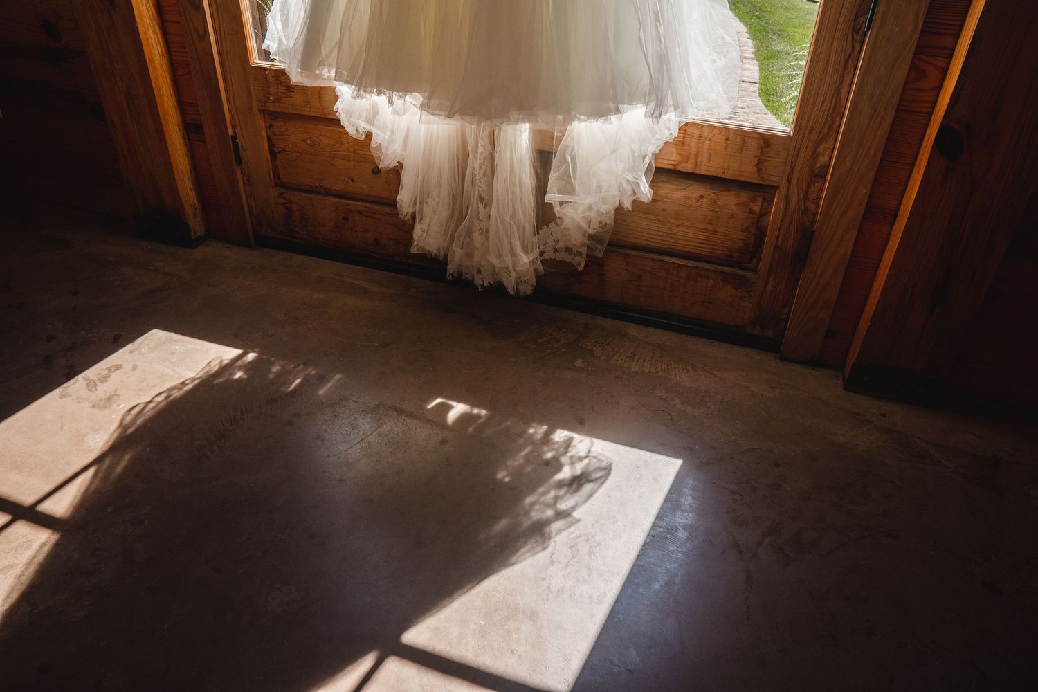 Wedding dress hanging in rustic barn doorway with sunlight casting lace shadows on concrete floor.