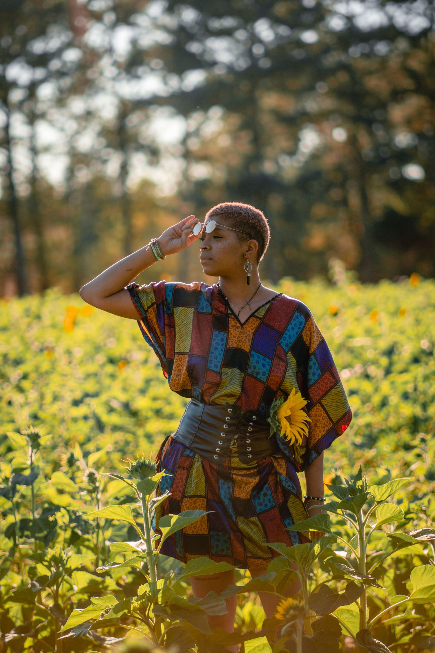 Woman in vibrant patchwork kaftan shielding eyes from sun while standing in sunflower field.