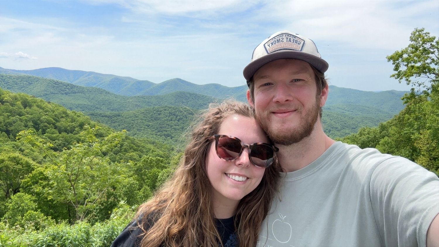 Smiling couple taking a selfie with a lush green mountain landscape stretching behind them.