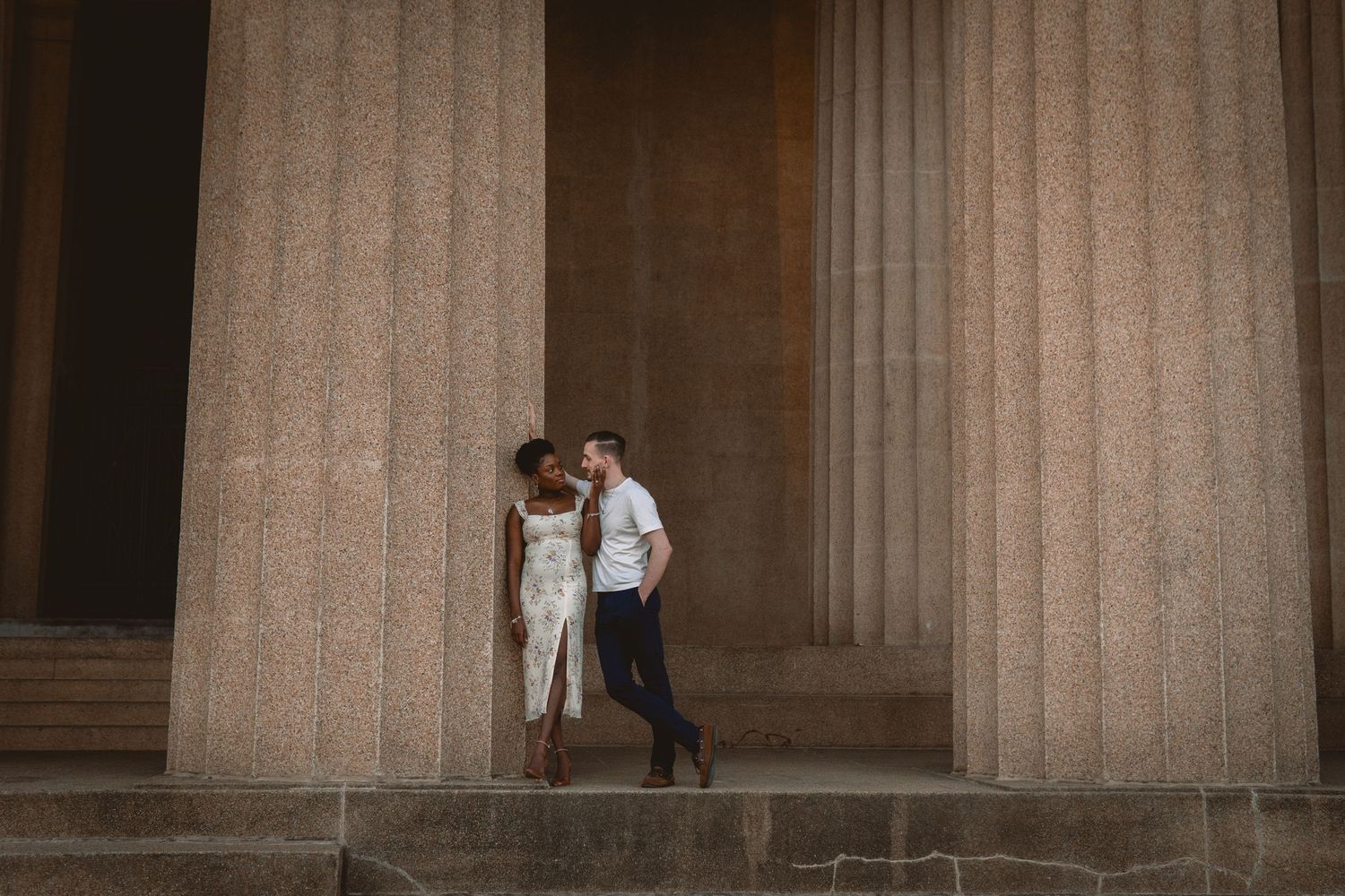 Interracial couple sharing an intimate moment between tall stone columns of a historic monument.