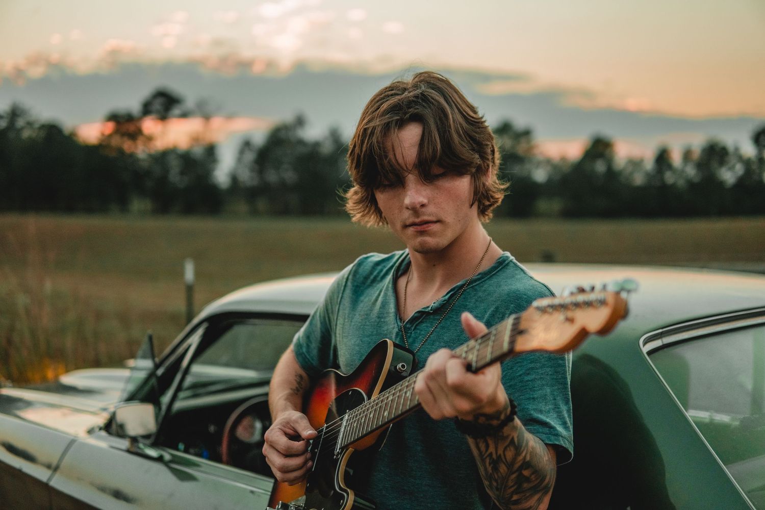 Young man with tattoos playing electric guitar leaning against vintage car at sunset.