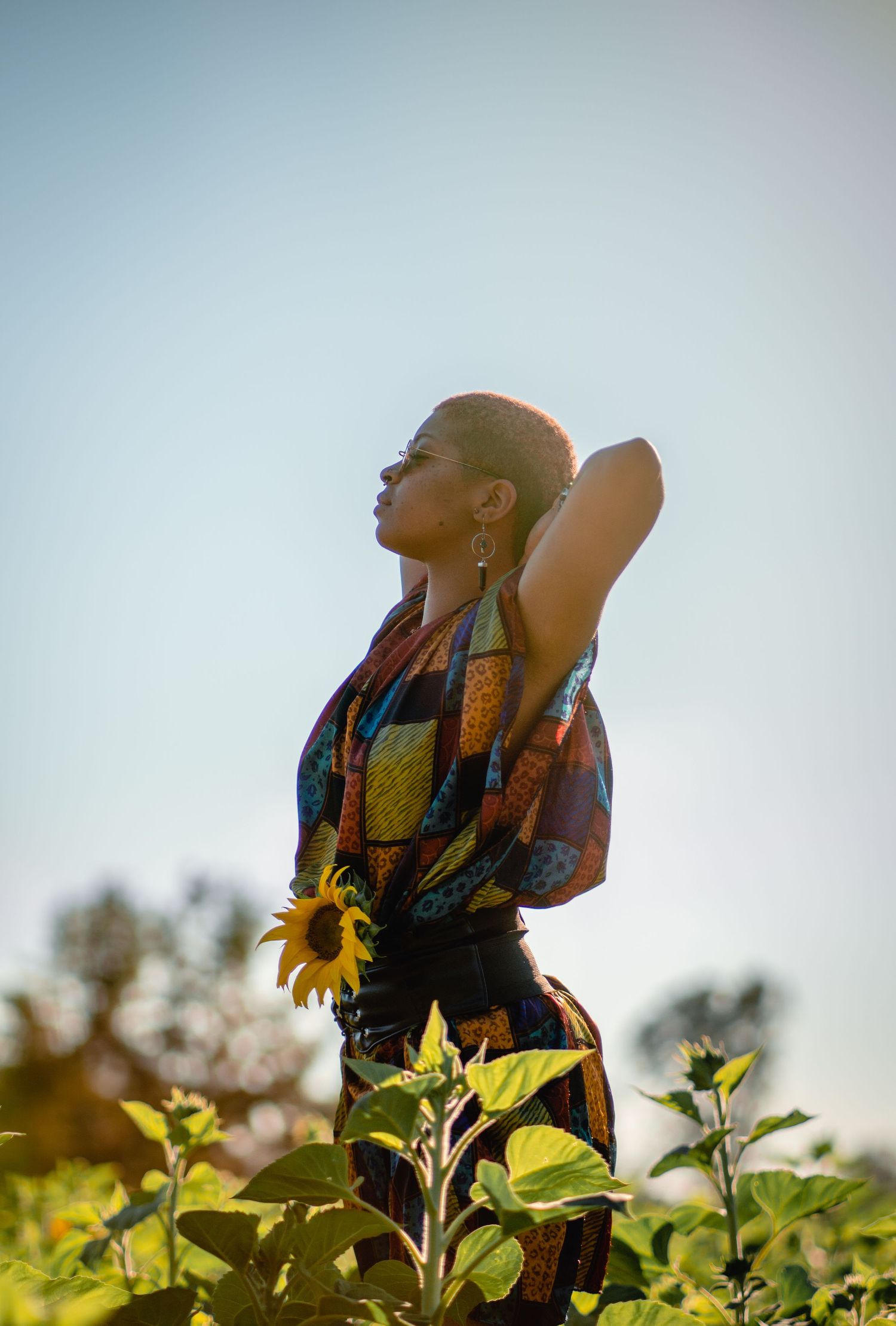 Woman in colorful African print dress holding sunflower while posing against clear sky in field.