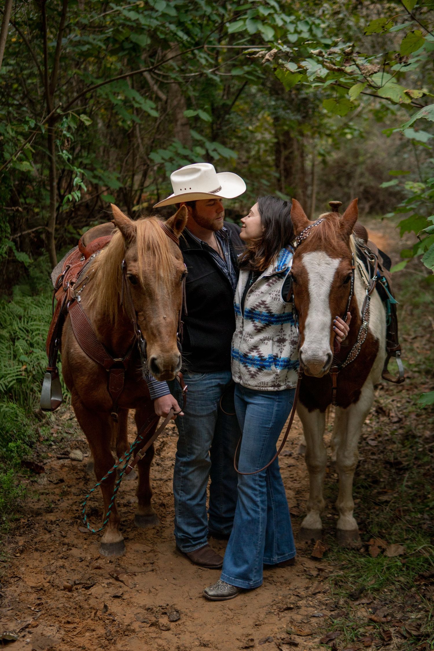 Couple standing close together with two saddled horses on a wooded forest trail.