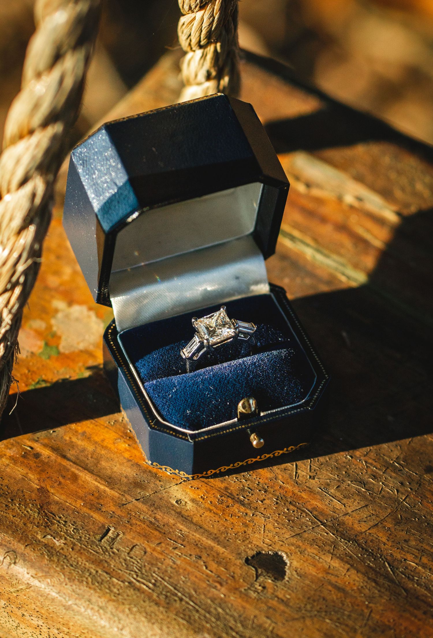 A princess-cut diamond engagement ring in a navy blue box displayed on a wooden rope swing.