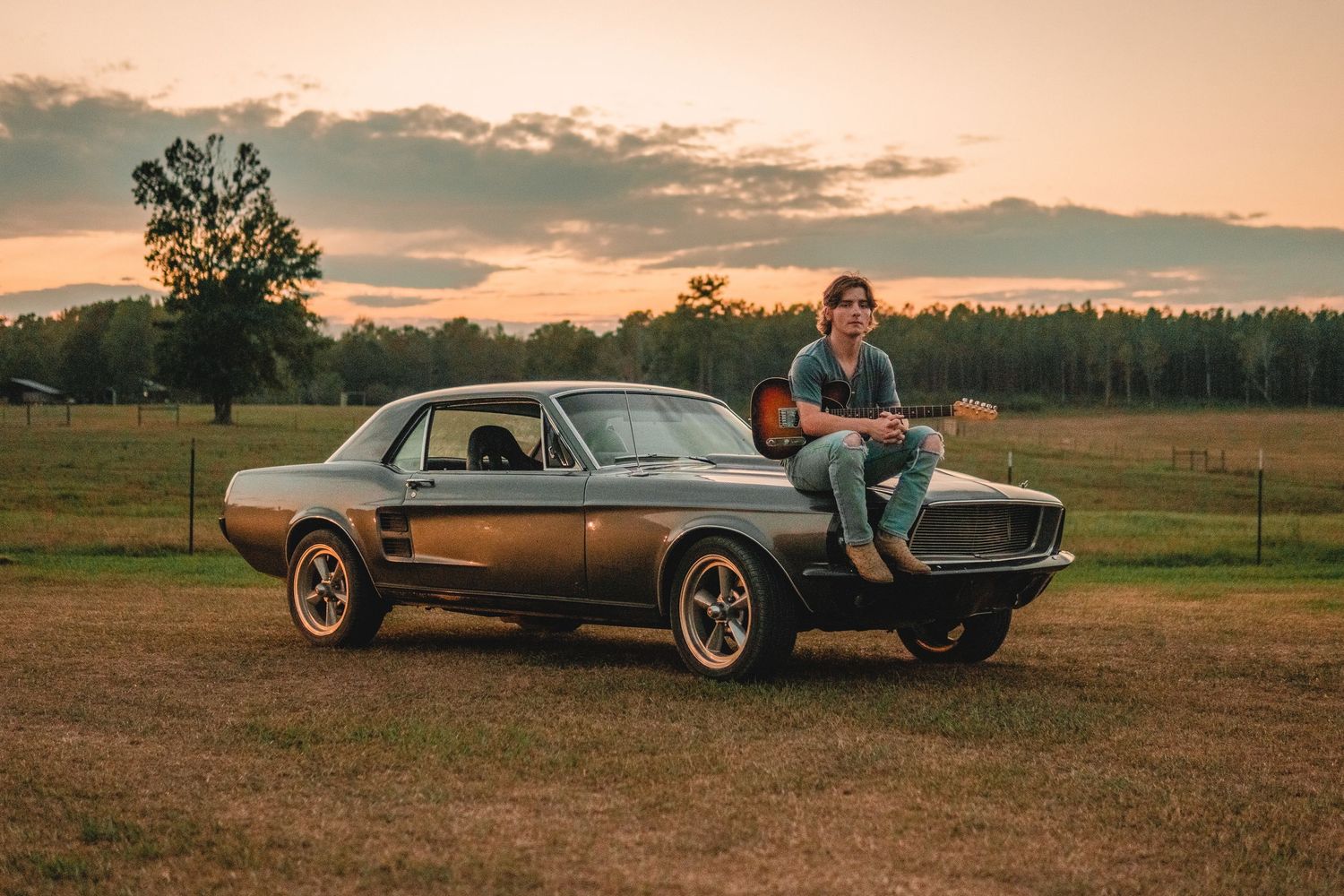 Young man sits on classic vintage Mustang holding guitar at sunset on rural farm.