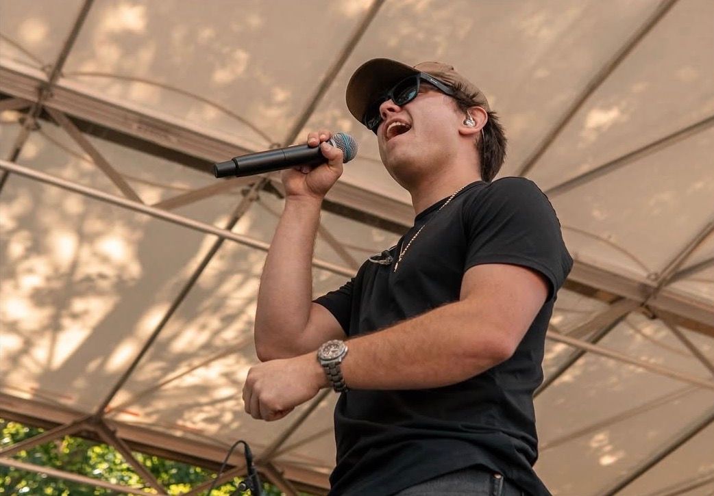 Male performer singing passionately into a microphone on an outdoor stage wearing a black shirt.