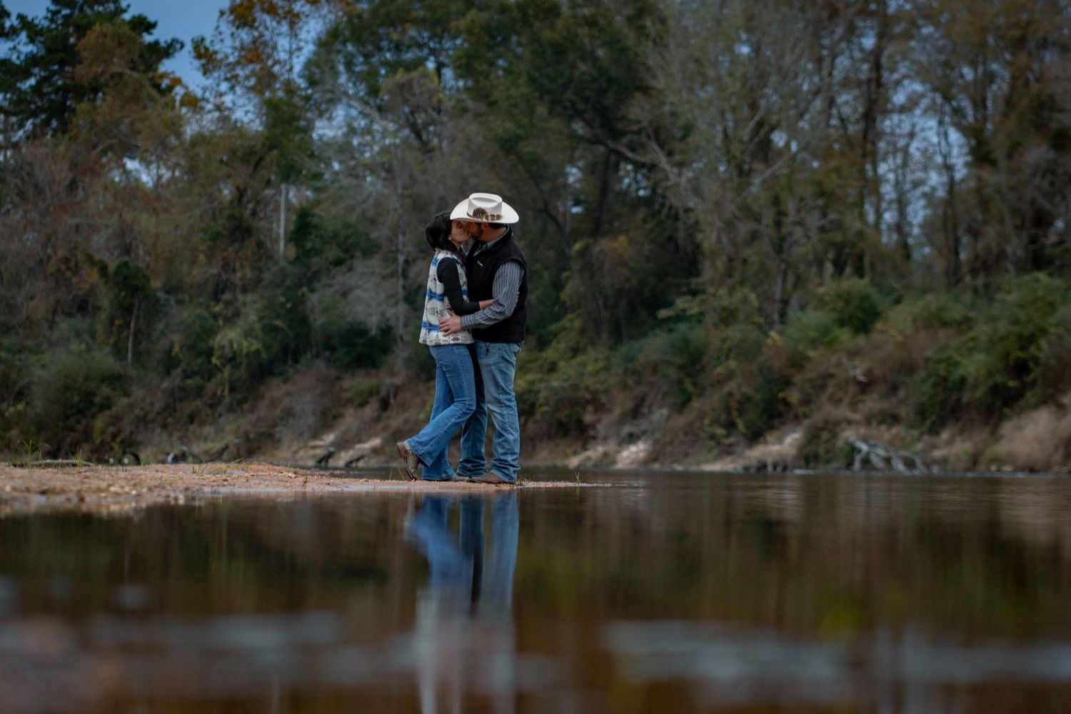 Couple sharing a romantic kiss while standing in a shallow river surrounded by lush green trees.