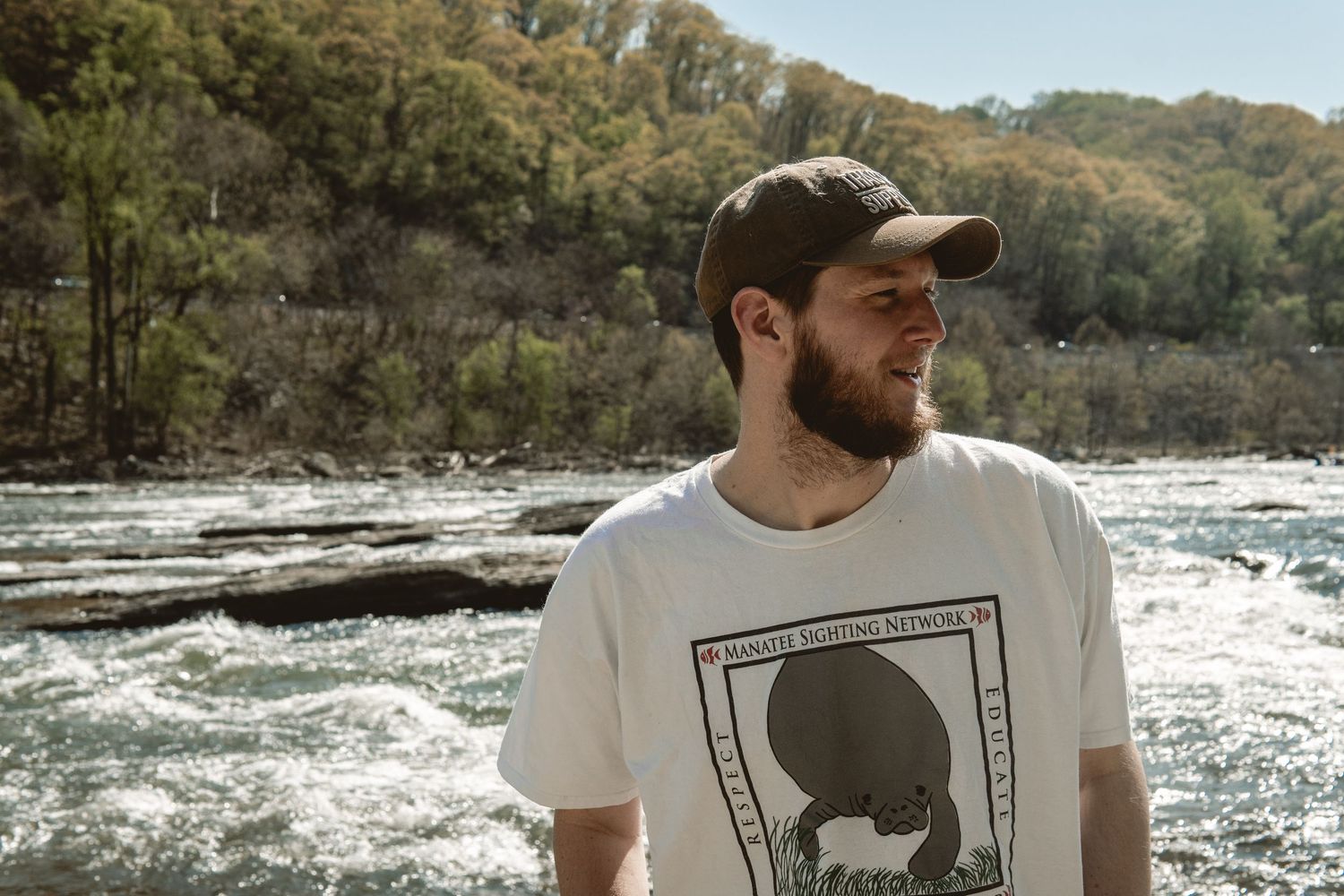 Bearded man in a white graphic tee gazing sideways near a rocky river with lush green trees behind.