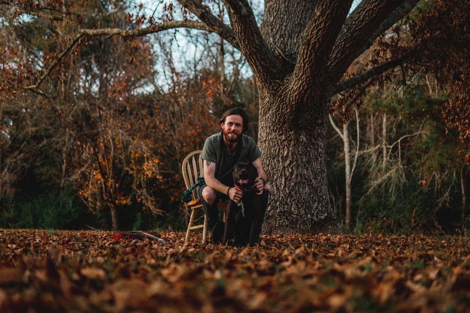 A bearded man smiles while sitting beside a large tree trunk surrounded by fallen autumn leaves.