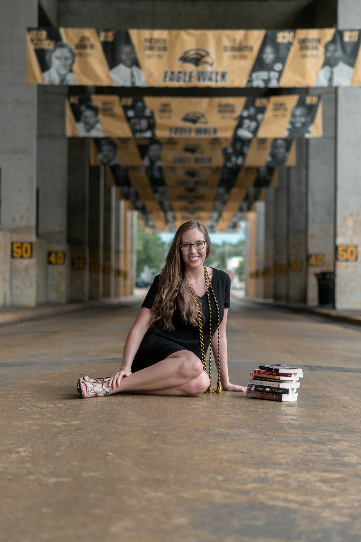 Young woman in black dress sitting cross-legged on stadium walkway with books and Eagle Walk banners.