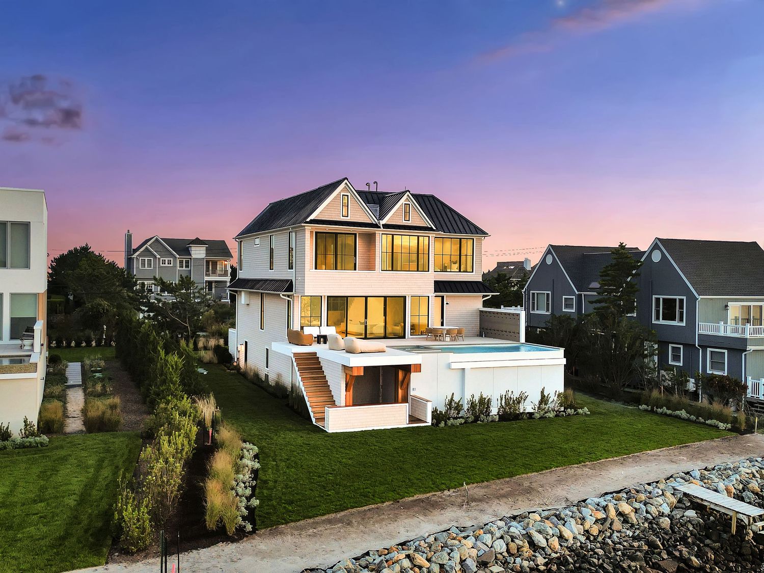 Luxury coastal home with white exterior, multiple balconies, infinity pool, and lush lawn at twilight.
