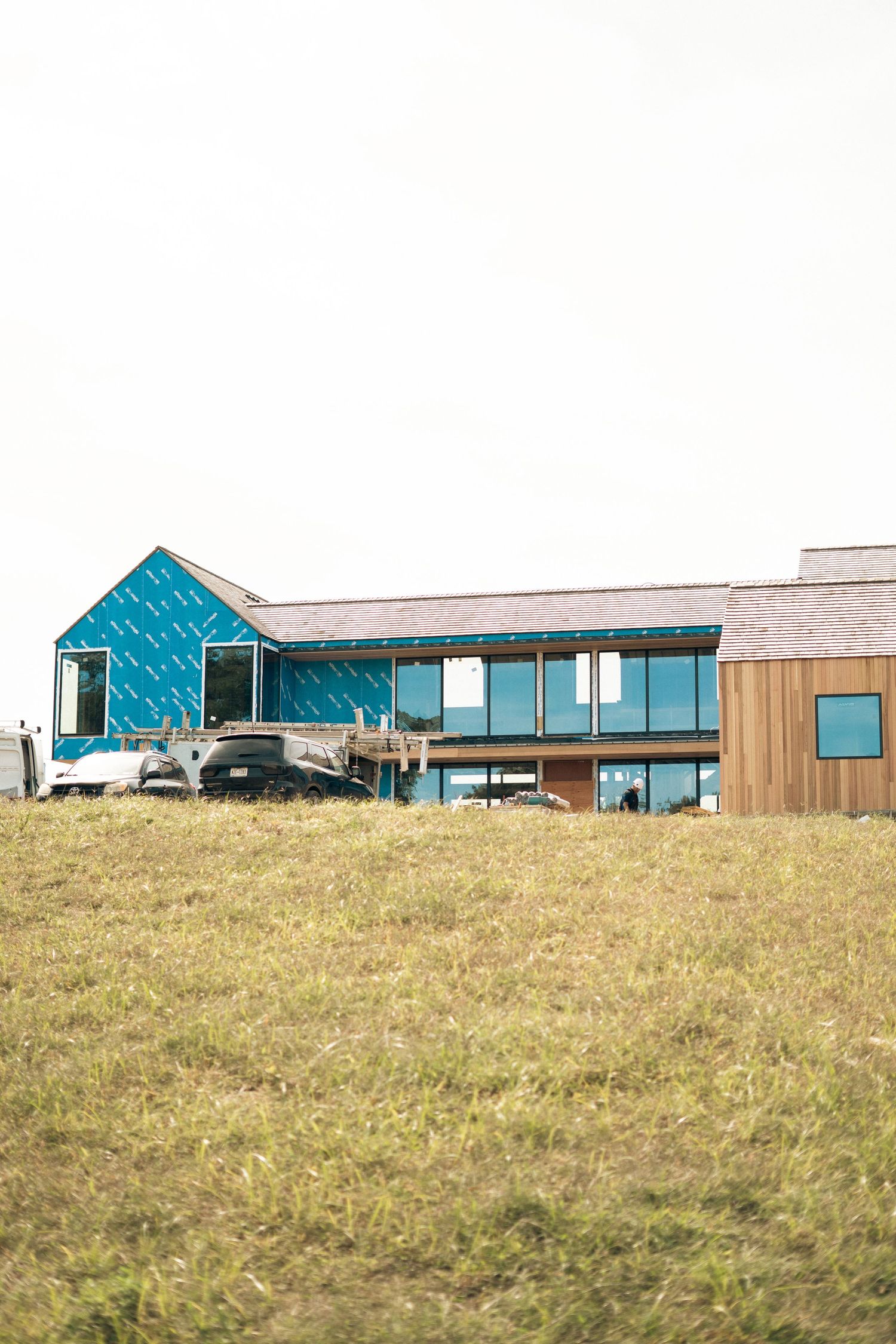 Modern home under construction with blue weather barrier and wood siding on a grassy hillside.