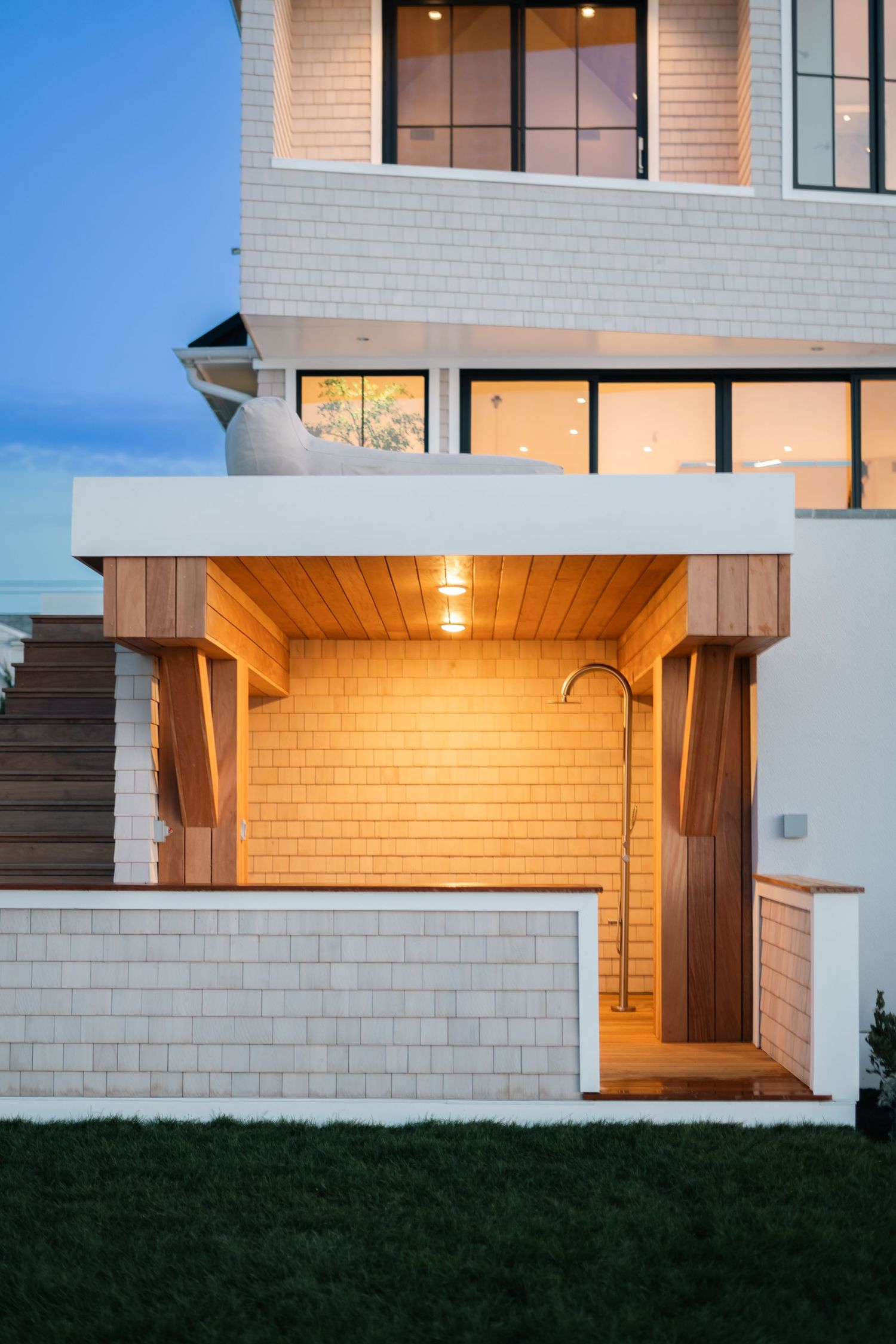 Modern coastal home exterior at dusk featuring cedar shingle siding, outdoor shower, warm lighting, and expansive windows.