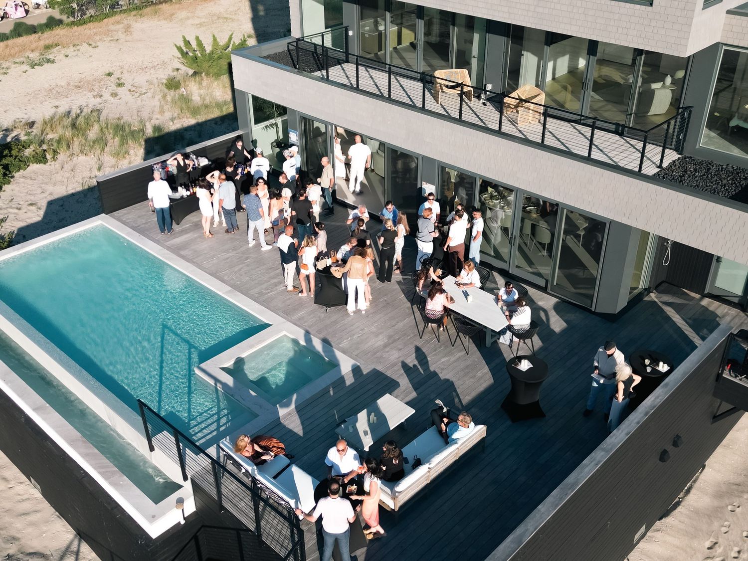 Aerial view of a modern beach house rooftop pool party with guests in white attire on a sleek deck.