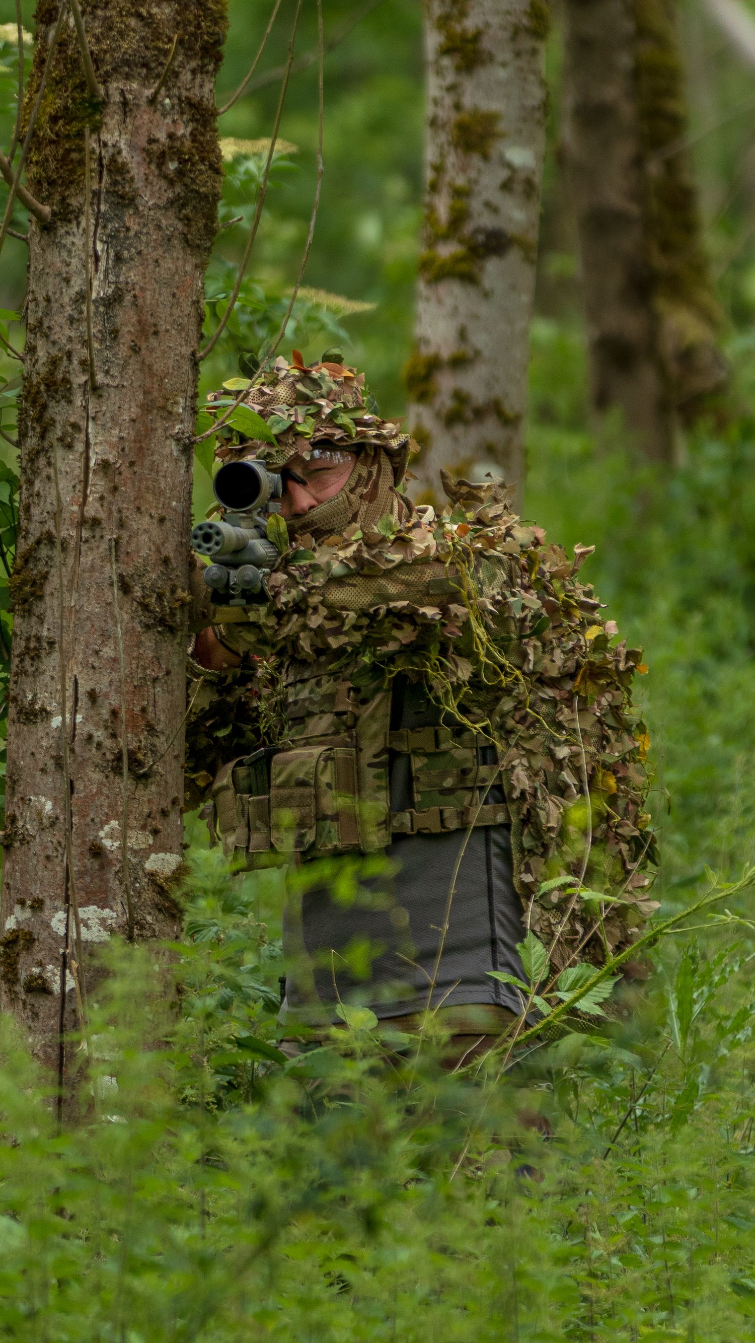 Camouflaged sniper in leaf ghillie suit aiming scoped rifle from behind tree in green woodland setting. Photo Taken by AJCoatesPhotography