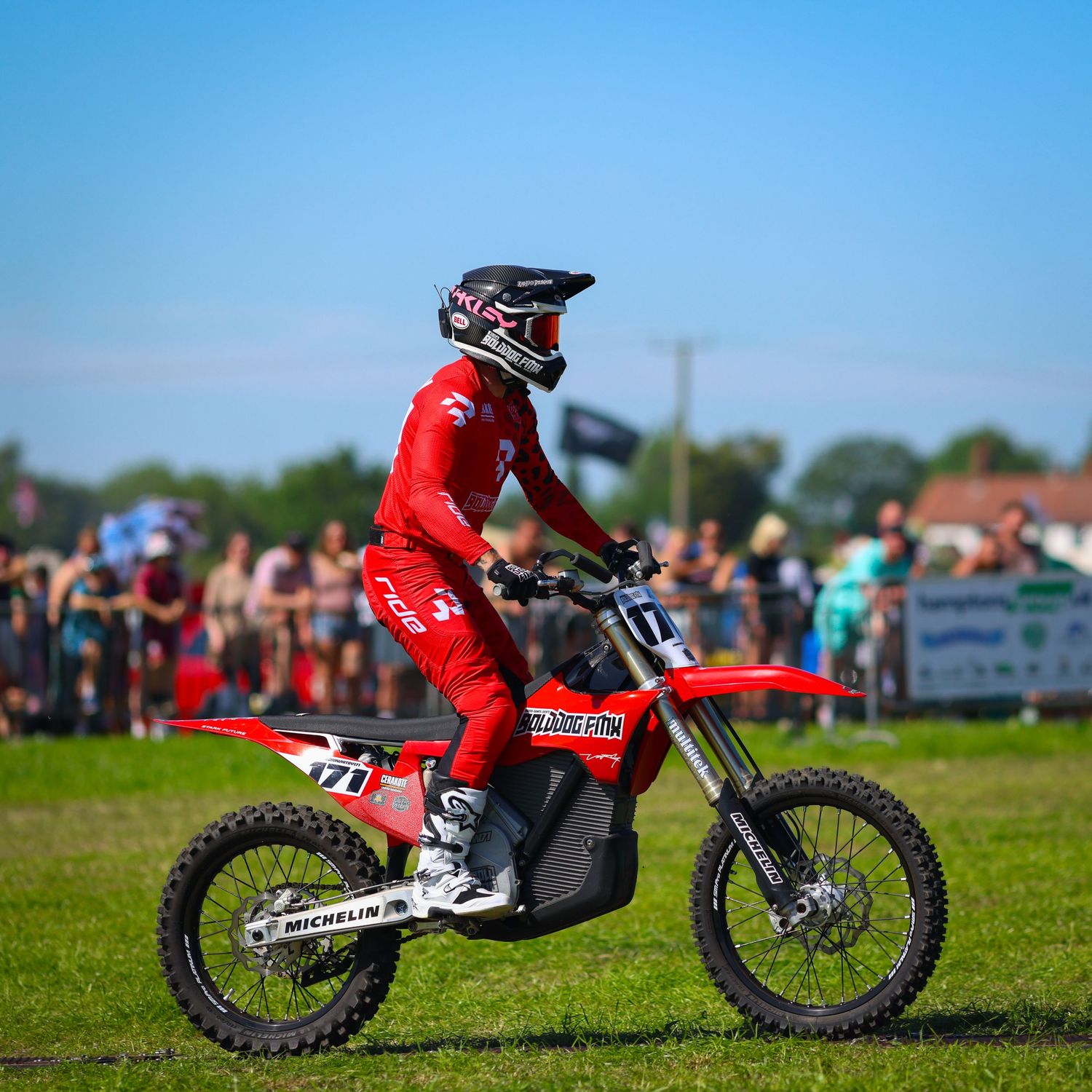 Motocross rider in red gear riding a Honda dirt bike on green grass with spectators in the background. Photo Taken by AJCoatesPhotography