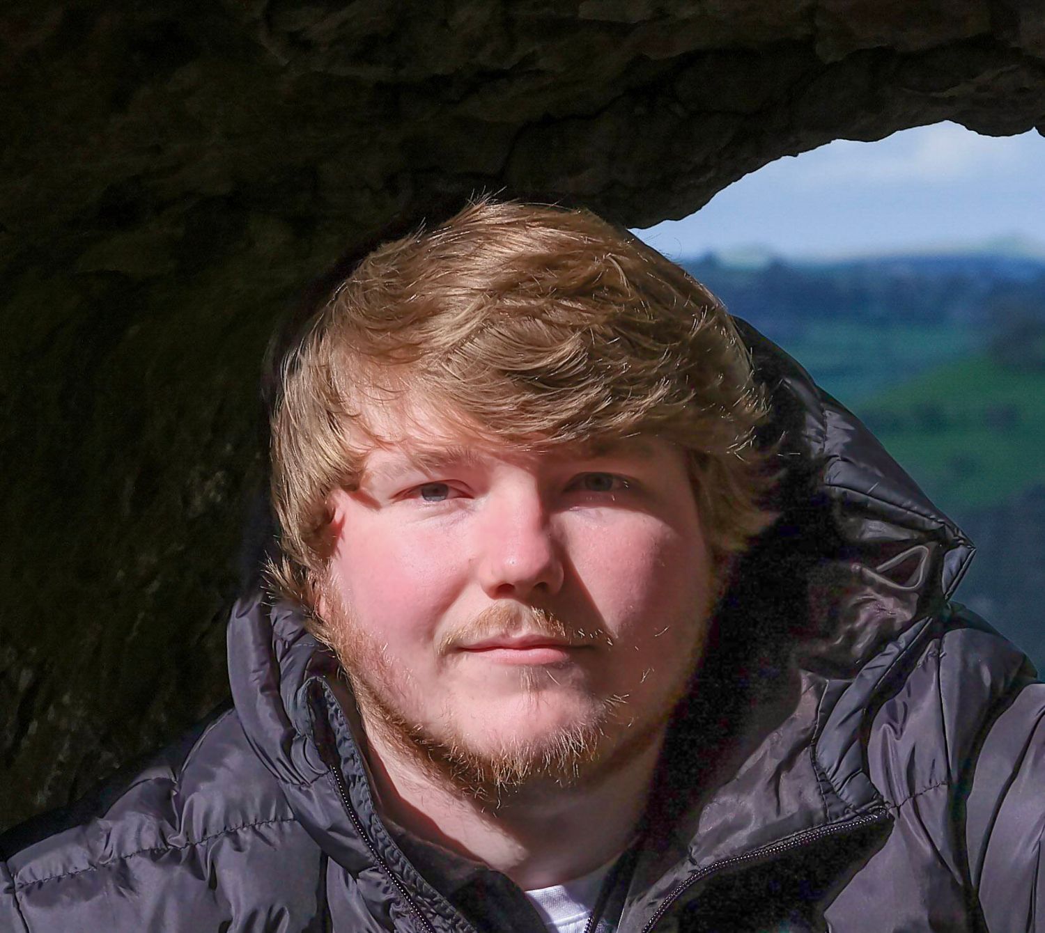 Young blonde man with stubble poses under a rock overhang with green countryside visible in the background. Photo Taken by AJCoatesPhotograp