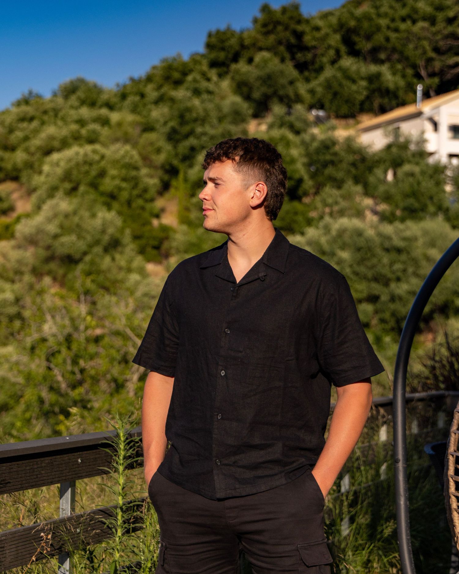 Young man in black short-sleeve shirt standing outdoors looking away thoughtfully. Photo Taken by AJCoatesPhotography