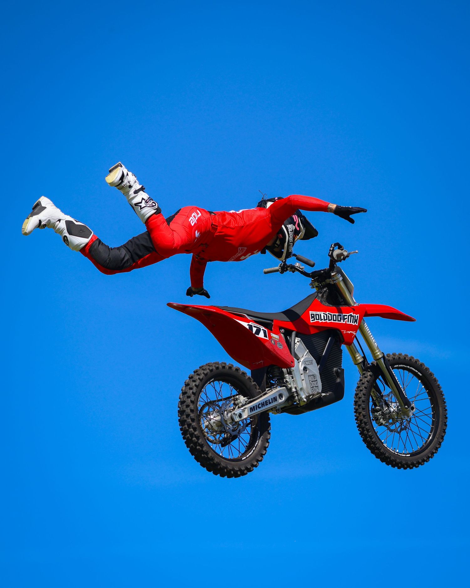 FMX rider in red suit performing a no-handed aerial stunt on a red dirt bike against blue sky. Photo Taken by AJCoatesPhotography