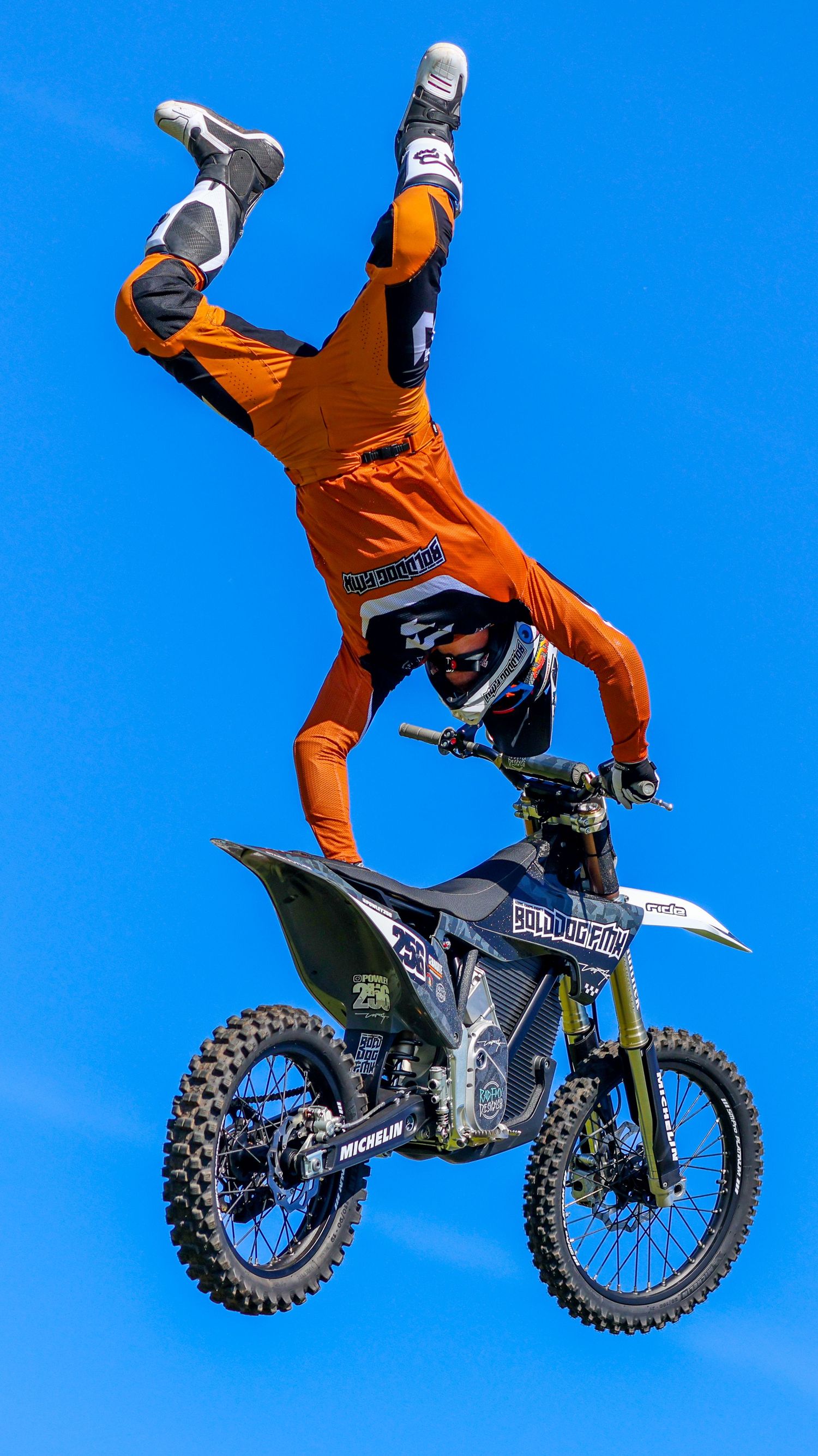 FMX rider in orange gear performs a headstand trick on a dirt bike against a clear blue sky. Photo Taken by AJCoatesPhotography