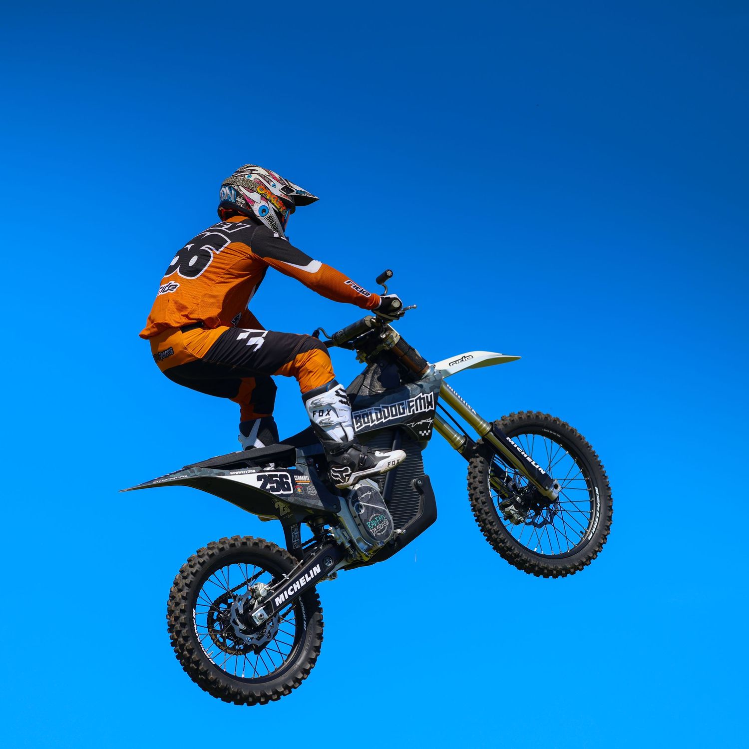 Motocross rider in orange gear jumping high on a black dirt bike against a vivid blue sky. Photo Taken by AJCoatesPhotography