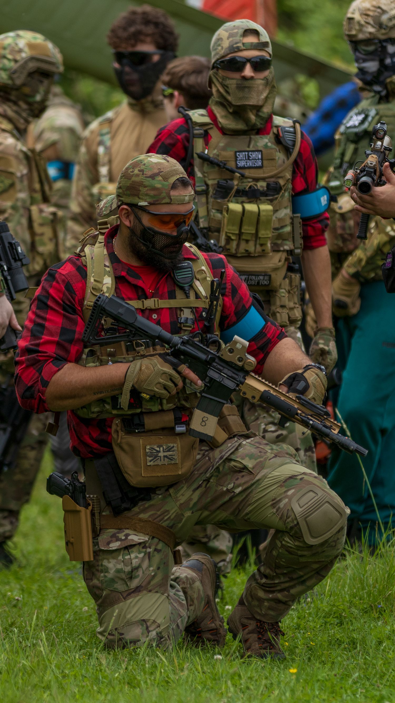 Airsoft players in tactical multicam gear and red plaid shirt kneeling with rifles on grass field. Photo Taken by AJCoatesPhotography