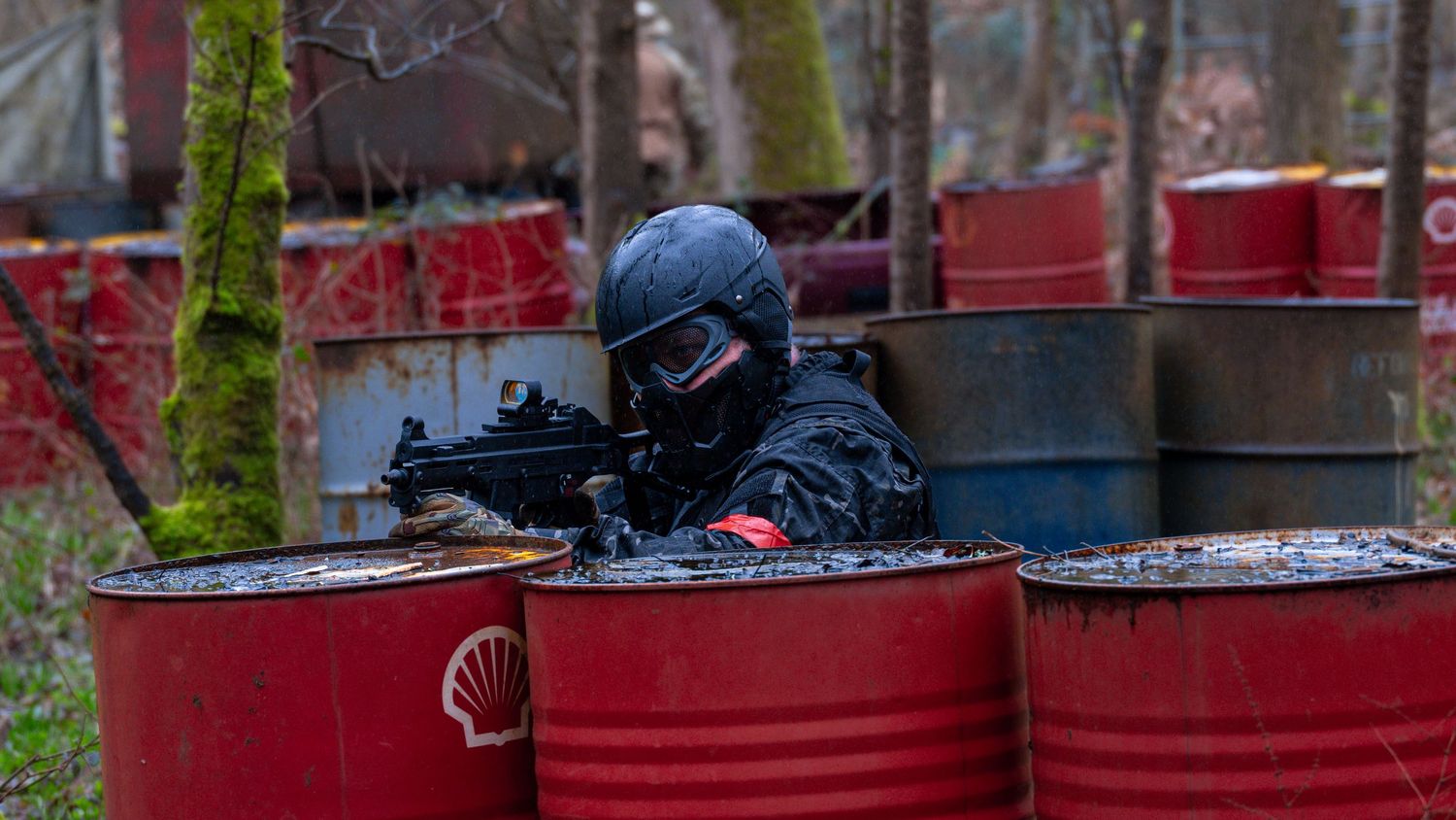 Airsoft Tactical operator in black helmet and gear aiming rifle behind red barrels in training scenario. Photo Taken by AJCoatesPhotography