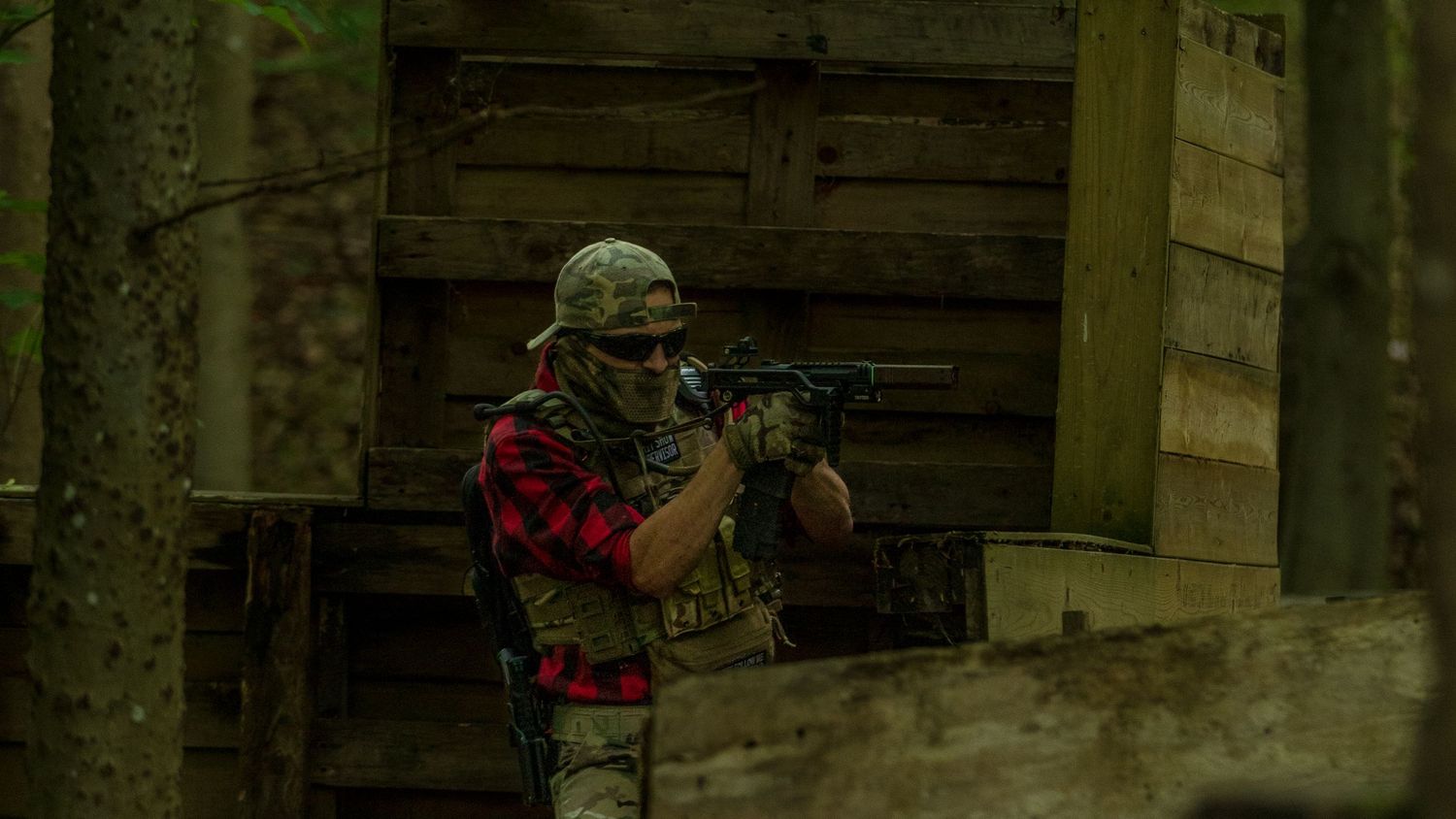 Airsoft player in red plaid shirt and tactical vest aiming rifle near wooden bunker in forest. Photo Taken by AJCoatesPhotography