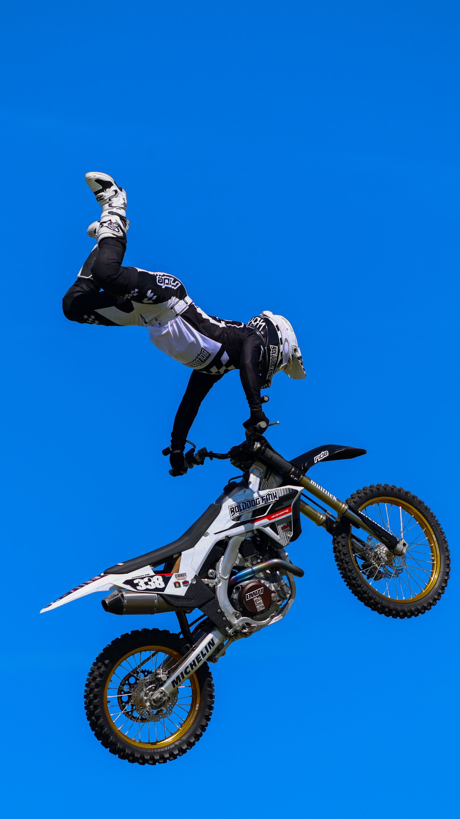 FMX athlete performing an inverted aerial trick on a white Husqvarna dirt bike against a vivid blue sky. Photo Taken by AJCoatesPhotography