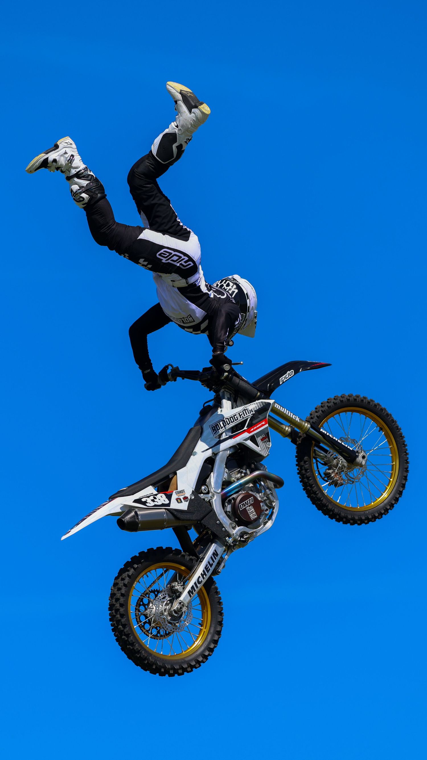 FMX rider in black and white gear executing an inverted vertical trick on a white and gold dirt bike. Photo Taken by AJCoatesPhotography