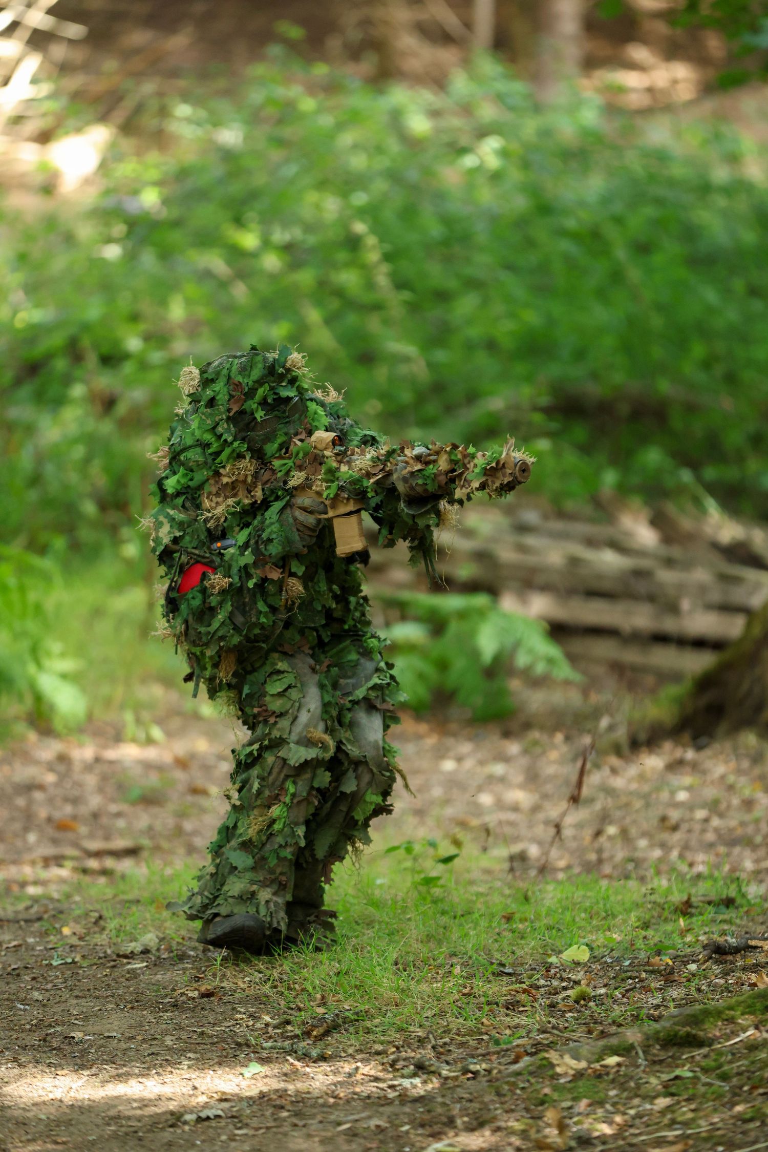 Person in ghillie suit camouflage aiming rifle in forest during military training exercise.