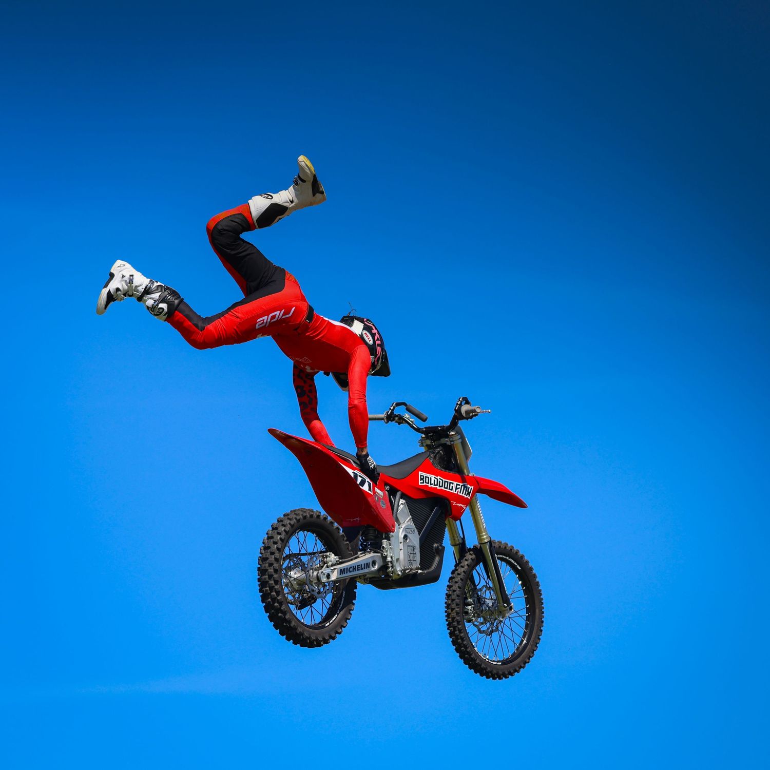 Daring FMX rider in red suit performs spectacular backflip stunt on dirt bike against blue sky. Photo Taken by AJCoatesPhotography