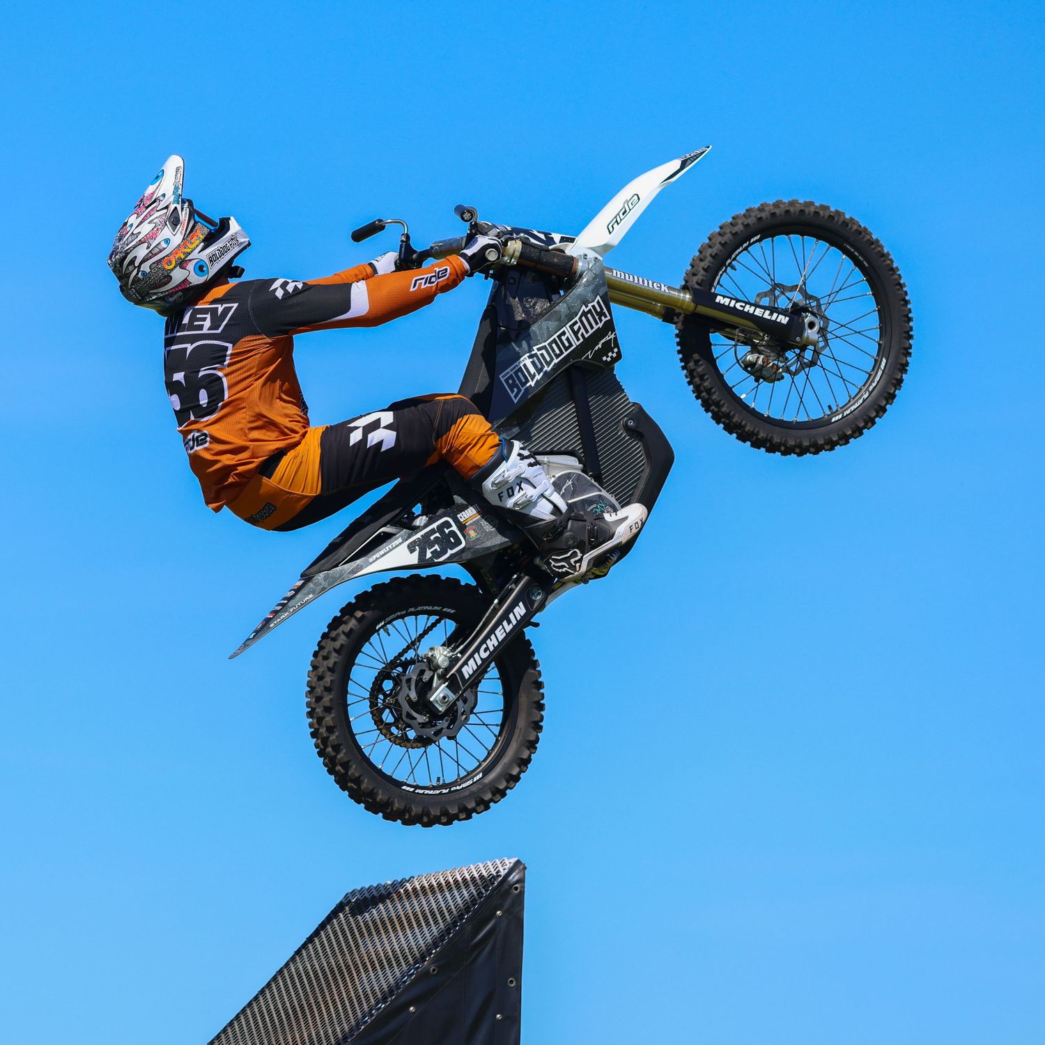 Motocross rider performing a dramatic aerial trick on an orange dirt bike against a clear blue sky. Photo Taken by AJCoatesPhotography